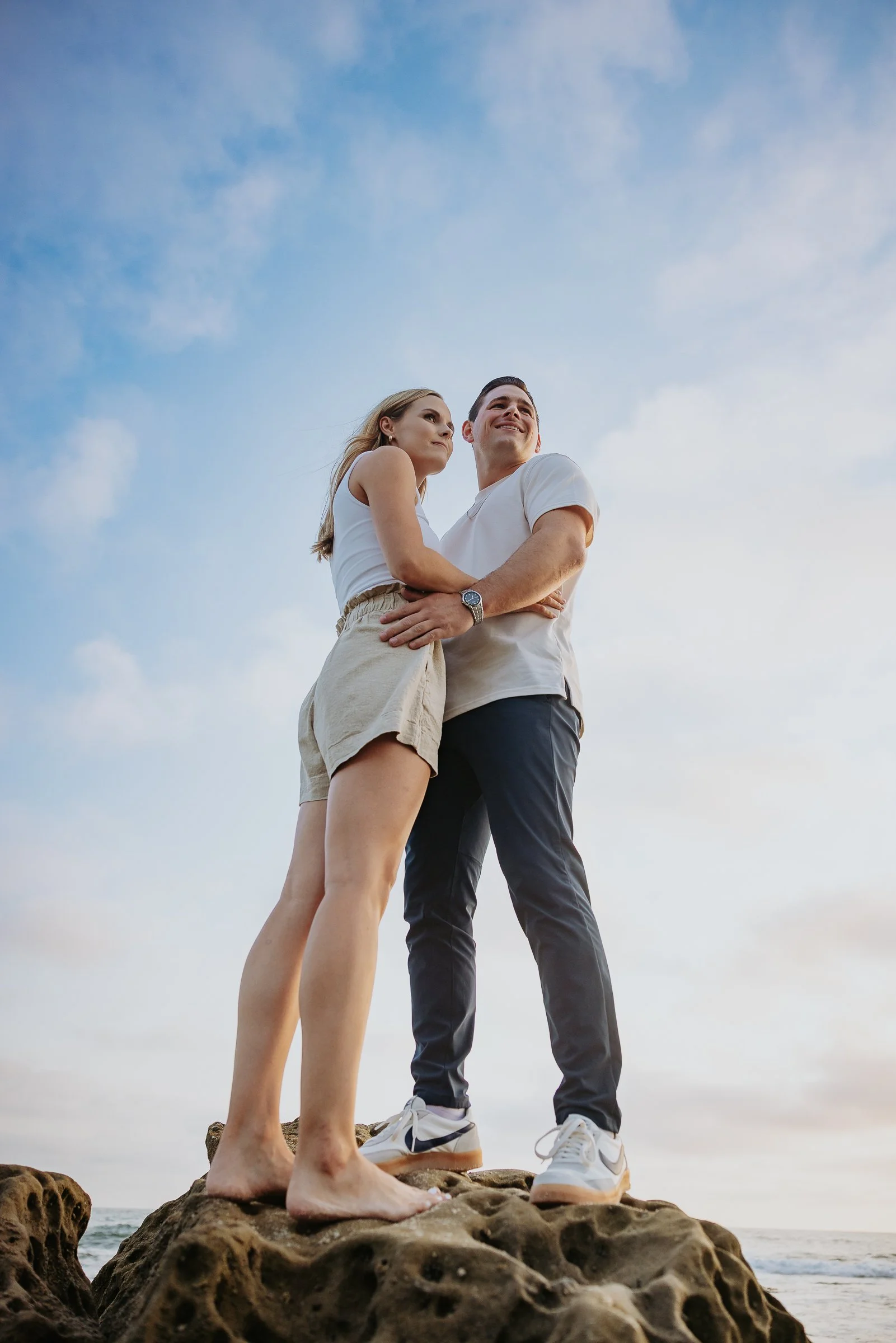 A couple standing on a rocky beach at sunset, with the woman in shorts and a tank top and the man in pants and a t-shirt, embracing each other and looking out at the ocean.