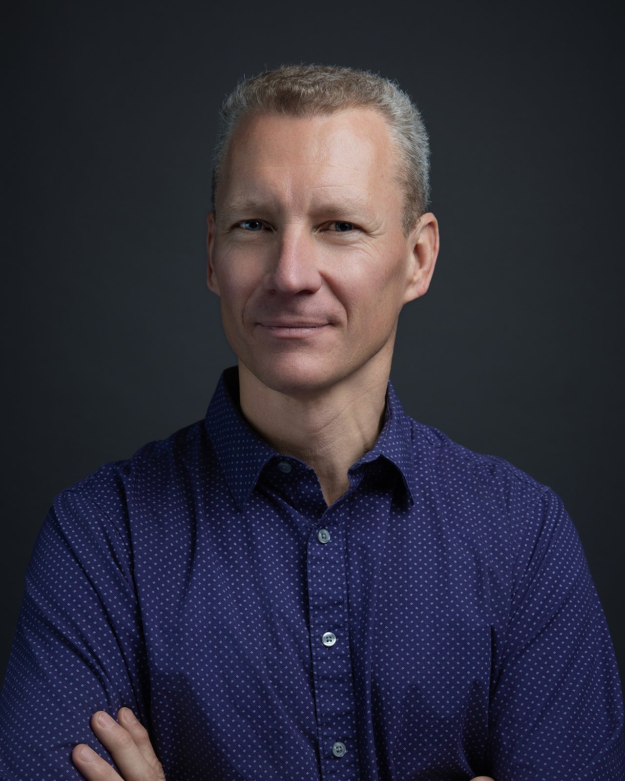 Portrait of a middle-aged man with short gray hair, wearing a dark blue dotted shirt, smiling slightly against a dark background.