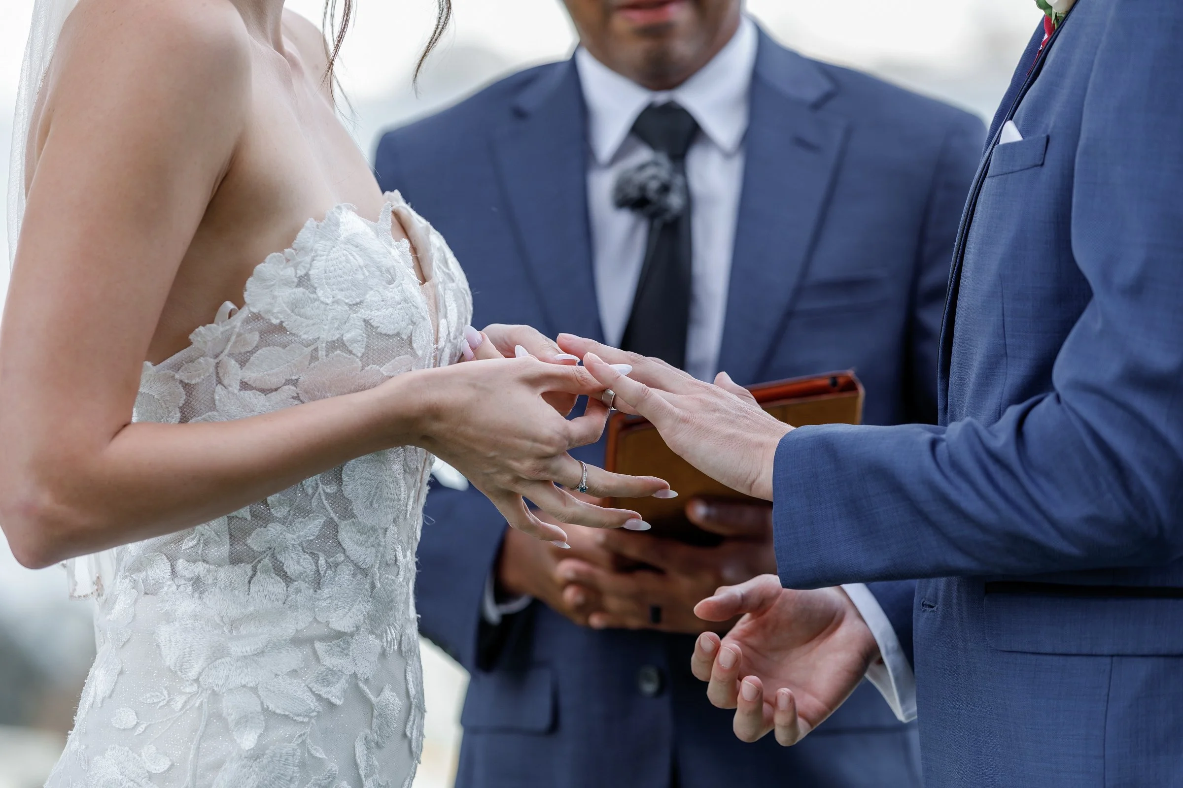 A bride and groom exchanging wedding rings during a ceremony, with an officiant holding a book in the background. The bride is wearing a lace wedding dress and the groom is wearing a blue suit.