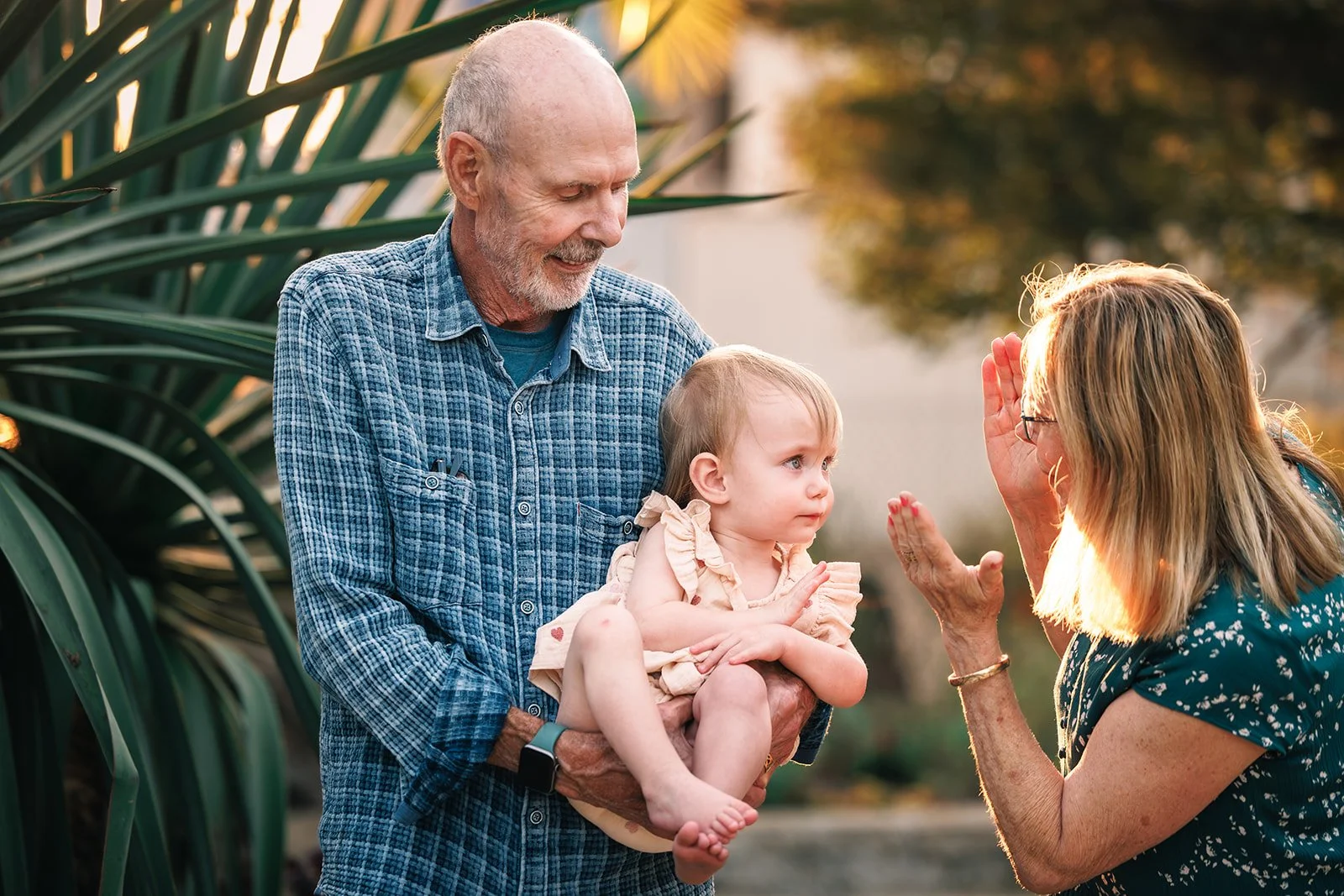 A little girl sits on her grandfather's arms as her grandmother raises her hand to her forehead in a gesture of greeting, outdoors during sunset.