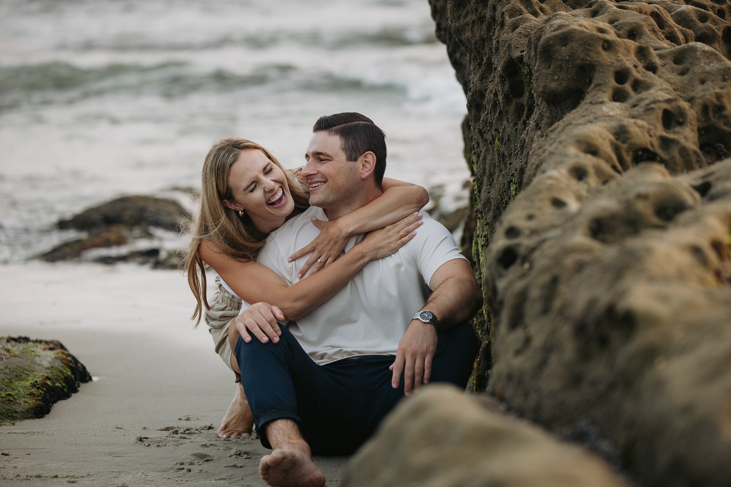A happy couple at the beach, sitting close together near rocks, smiling and laughing.