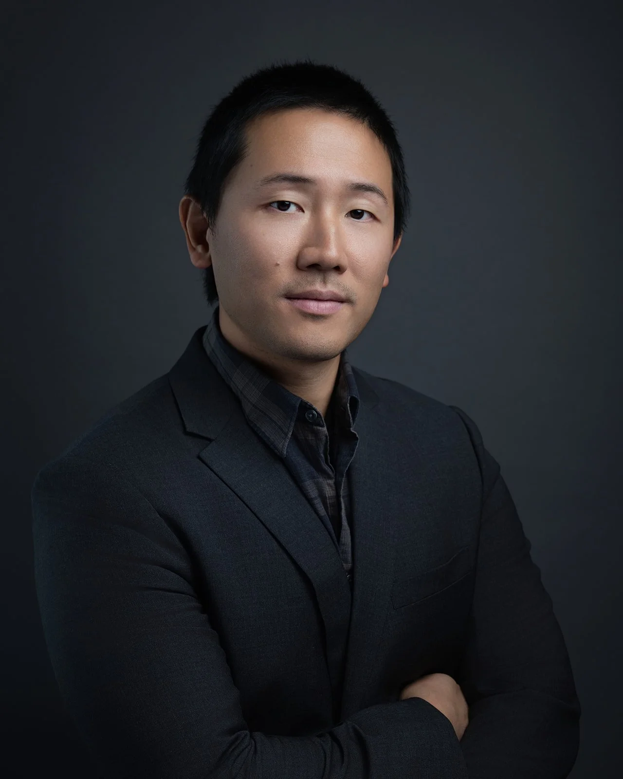 Portrait of a man with short black hair wearing a black suit jacket and a dark shirt, standing with arms crossed against a dark background.