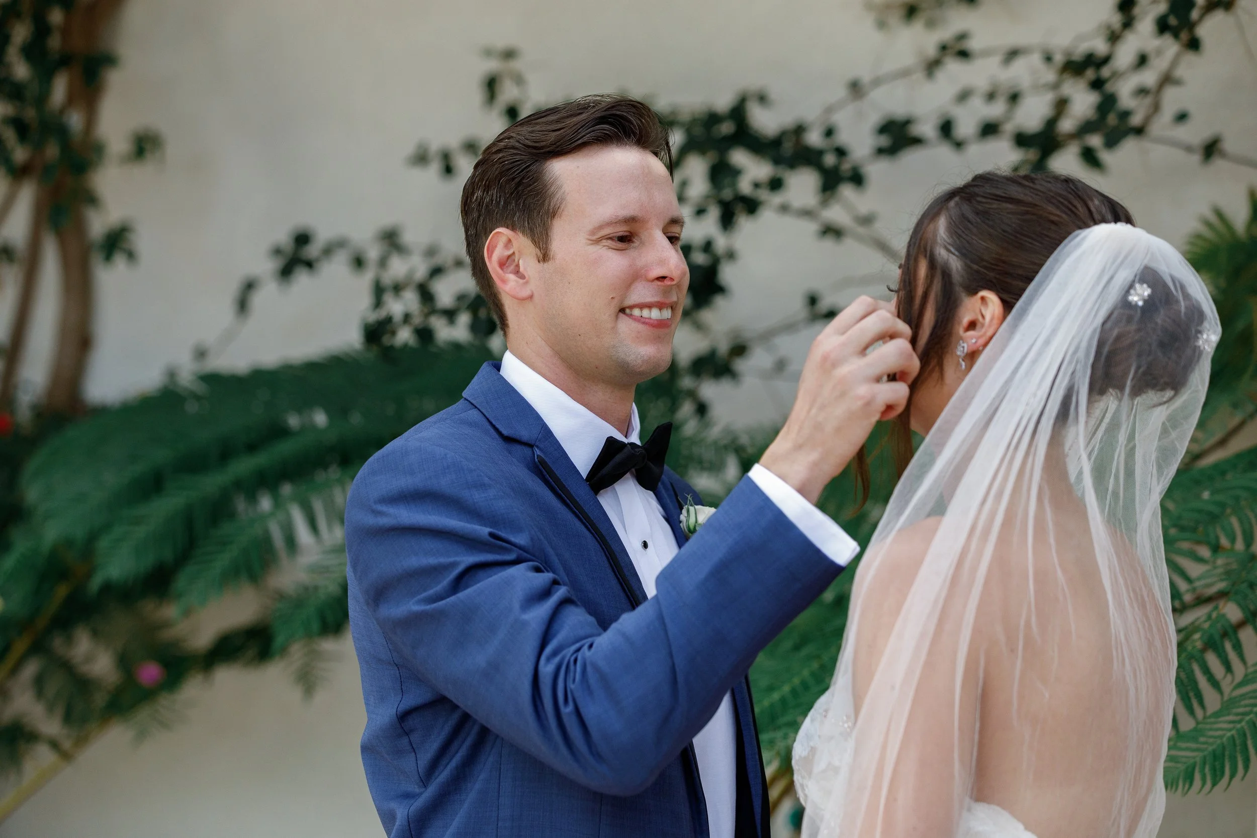 A groom in a blue tuxedo gently touches a bride's face during their wedding ceremony. The bride wears a veil and earrings, with a blurred green plant background.