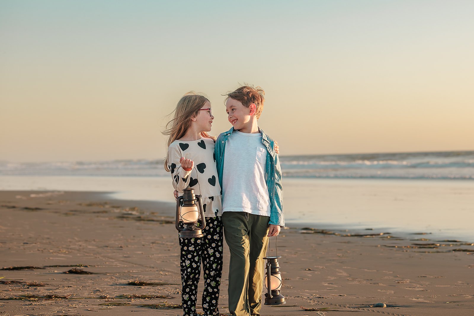 A young boy and girl walking on the beach during sunset, smiling at each other, each holding lanterns.