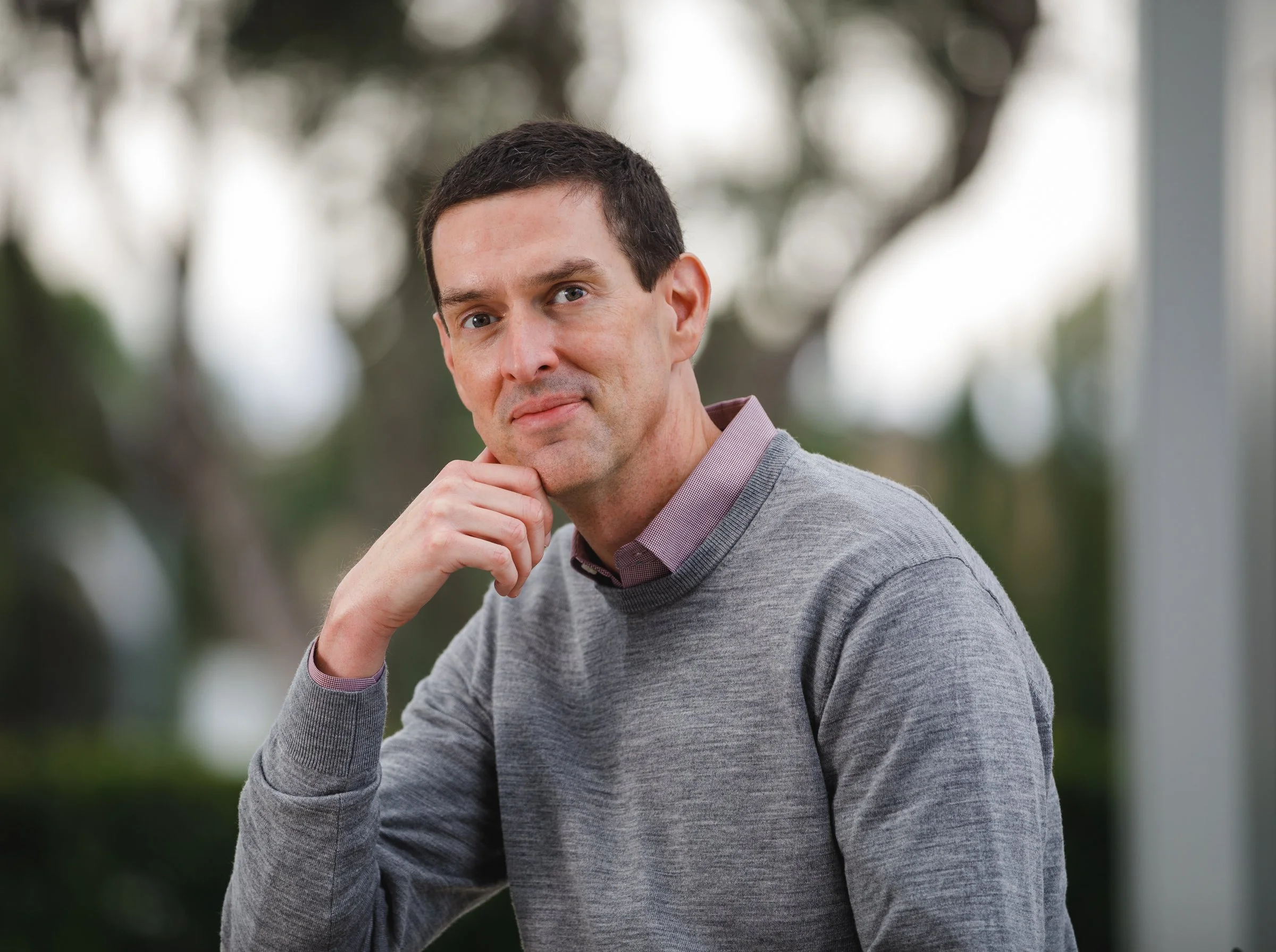 Portrait of a middle-aged man with dark hair, wearing a gray sweater over a collared shirt, sitting outdoors with a blurred natural background.