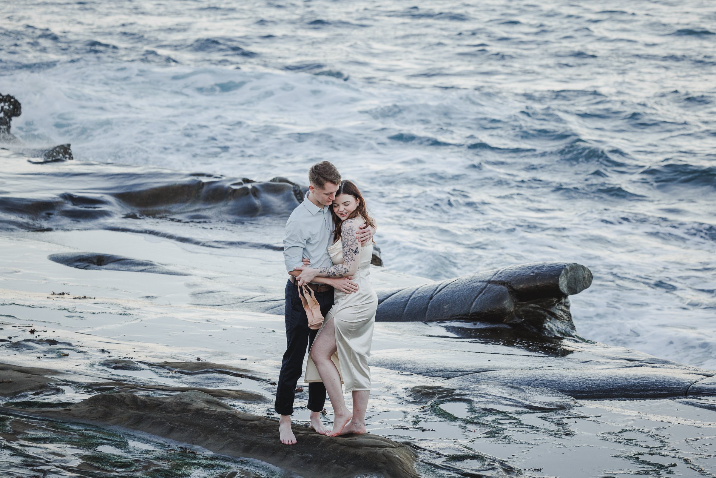 A couple hugging on a rocky beach with the ocean waves crashing behind them.