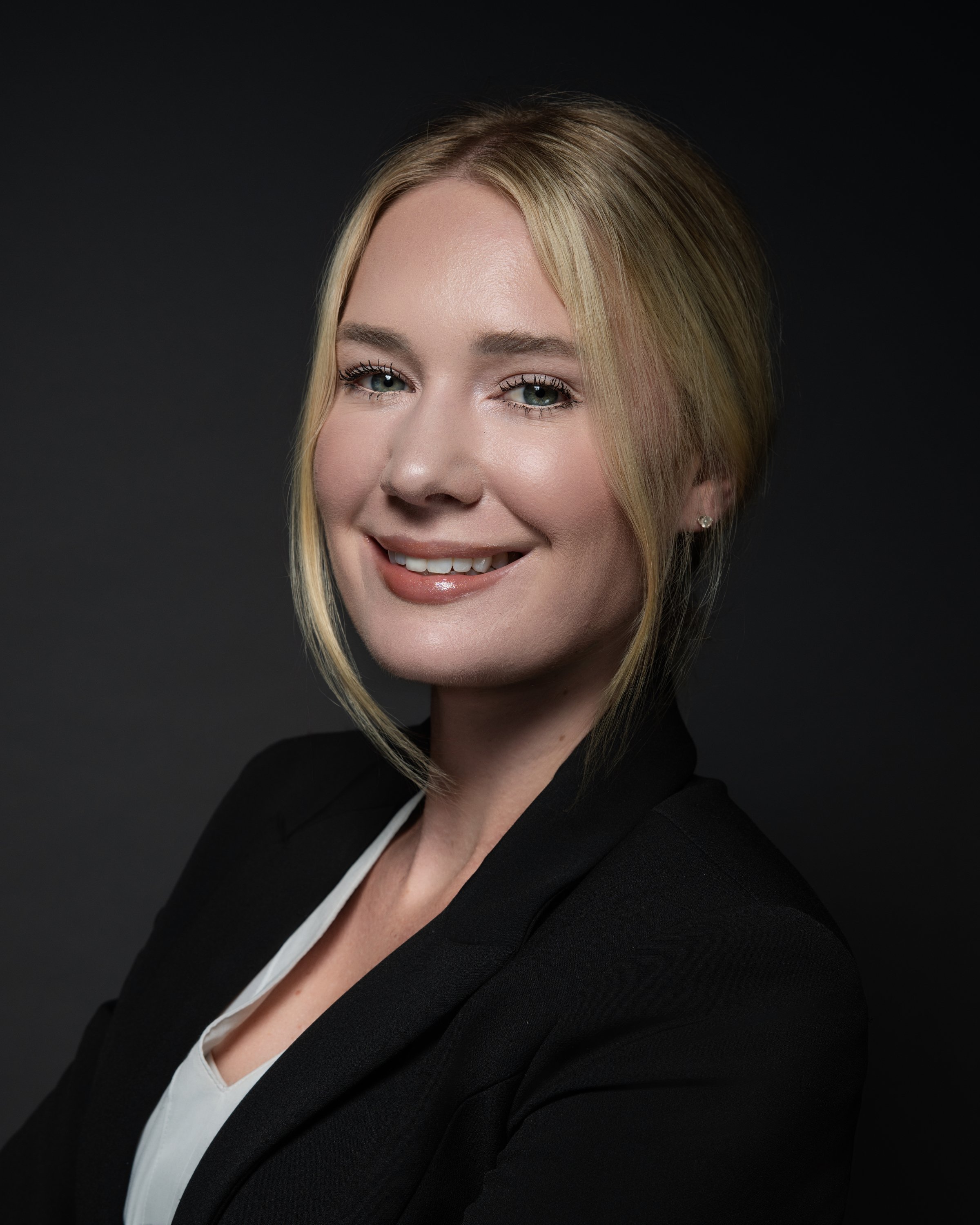A professional woman with blonde hair, wearing a black blazer and a white blouse, smiling in front of a dark background.