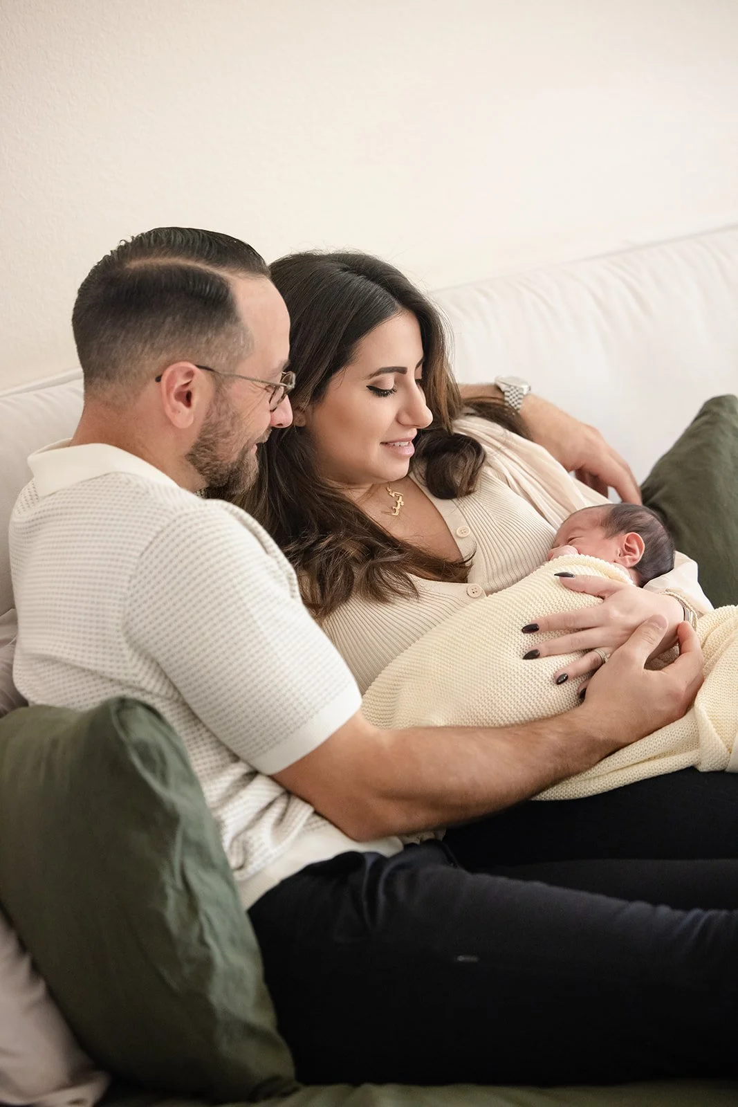 A family sitting on a sofa, with a woman holding a newborn baby and a man leaning close to her, all looking at the baby lovingly.