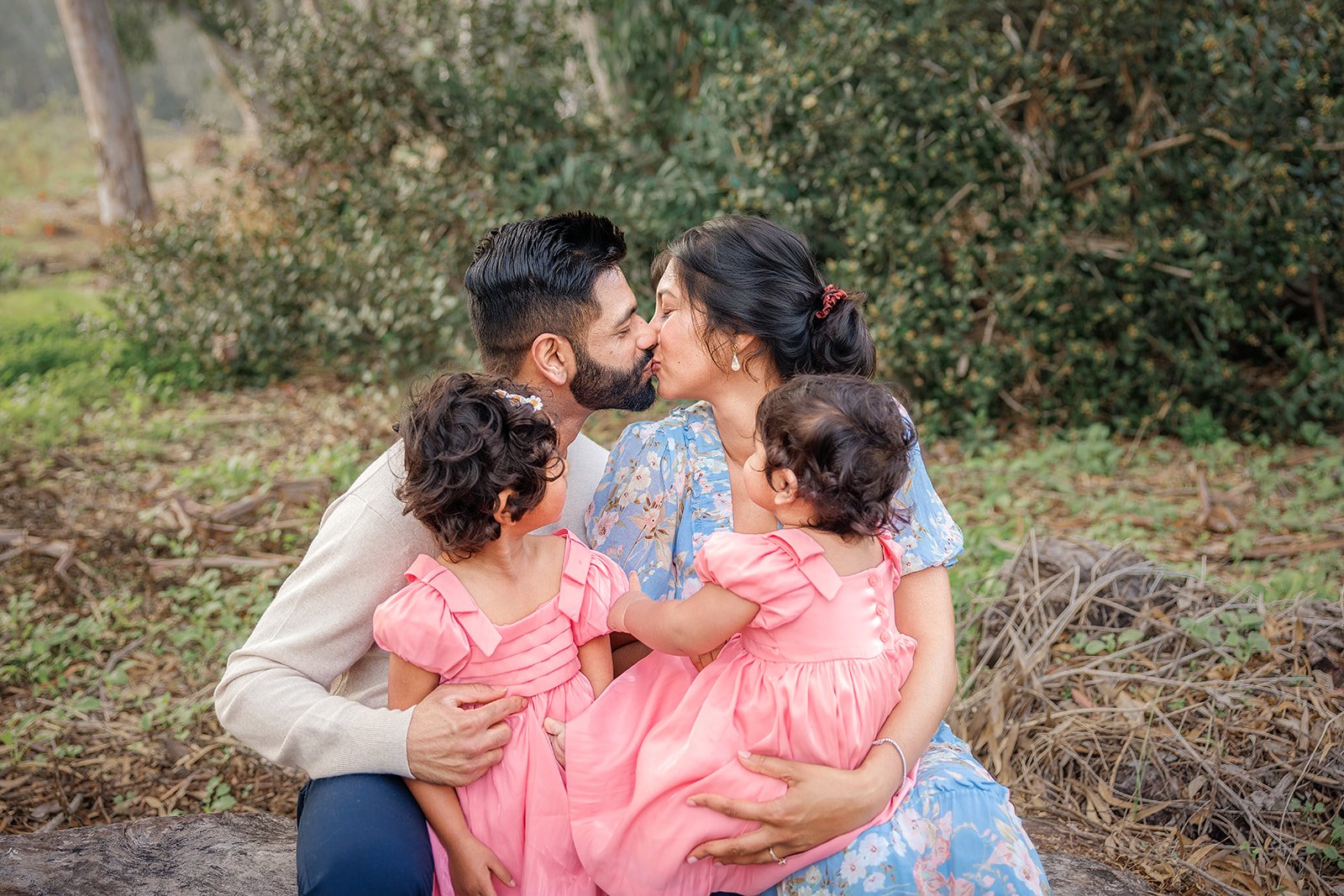 A family of four sitting outdoors, kissing and embracing each other, surrounded by greenery and trees.