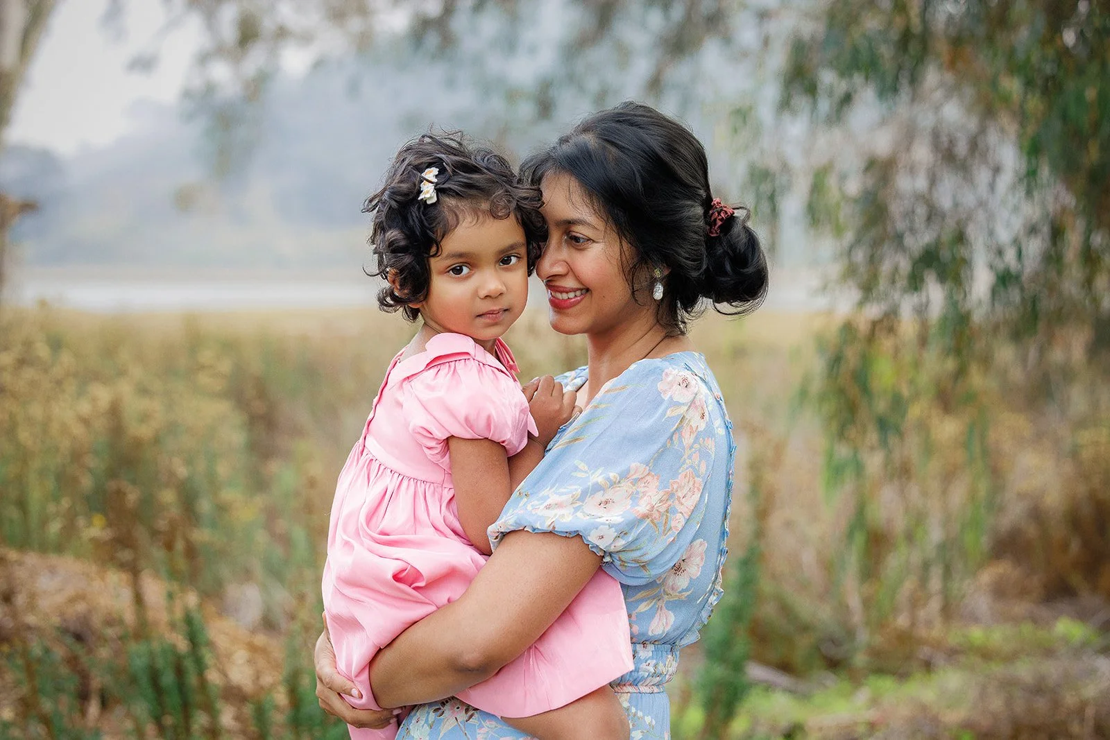 A woman holds a young girl outdoors near a body of water, with trees and foliage in the background. The woman is smiling and wearing a light blue floral dress, while the girl is wearing a pink dress with puffed sleeves. The girl has curly hair decora