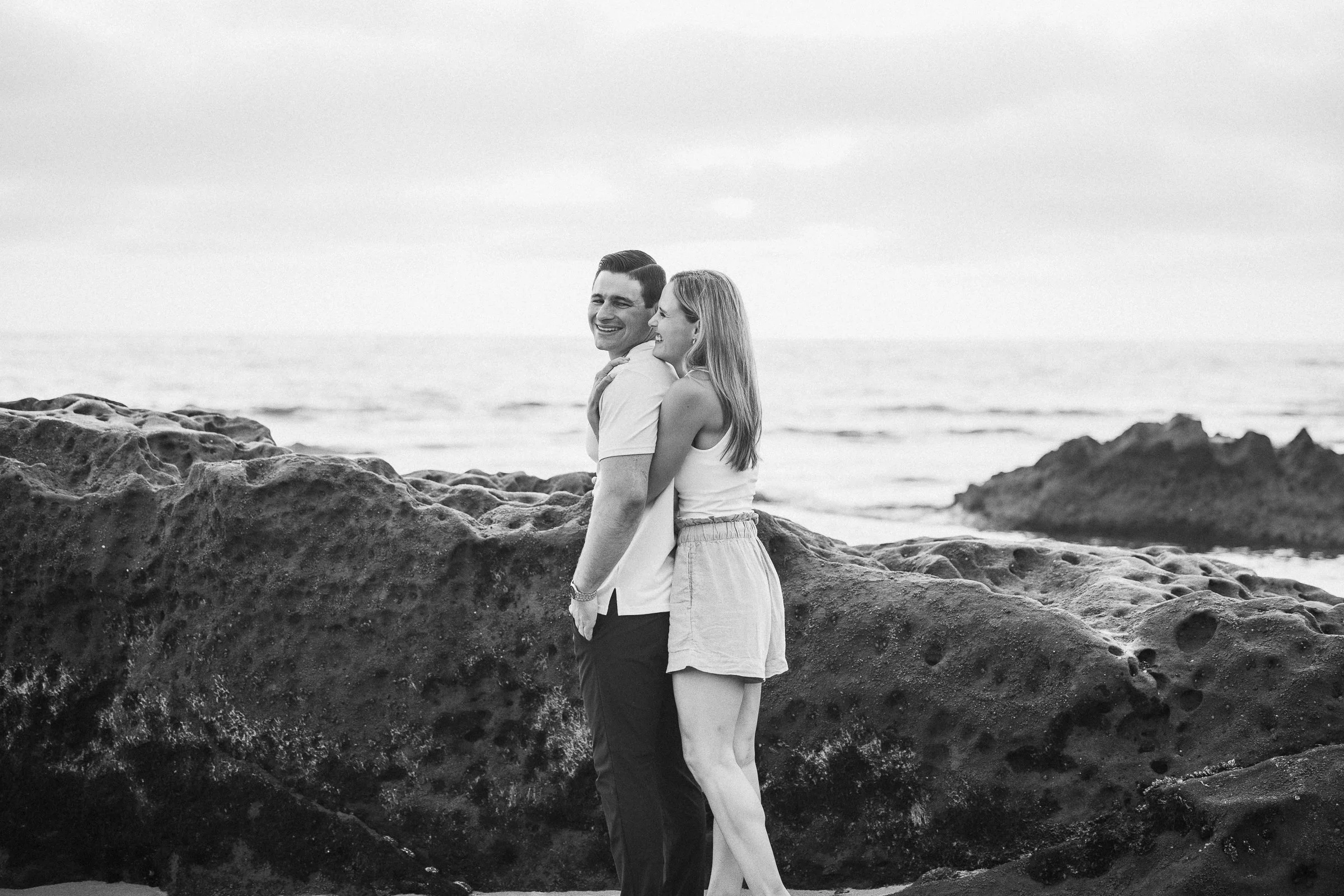 A smiling couple stands on a rocky beach near the ocean, embracing each other.