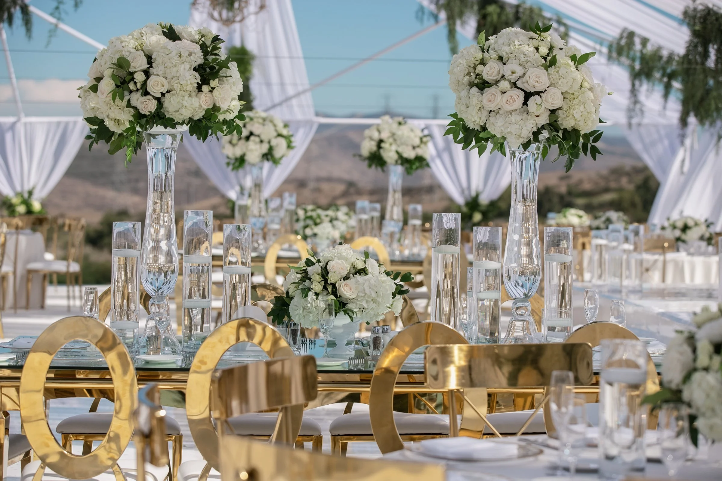 Elegant outdoor wedding reception setup with tall floral centerpieces, gold chairs, and glass tableware under a white tent with scenic mountains in the background.