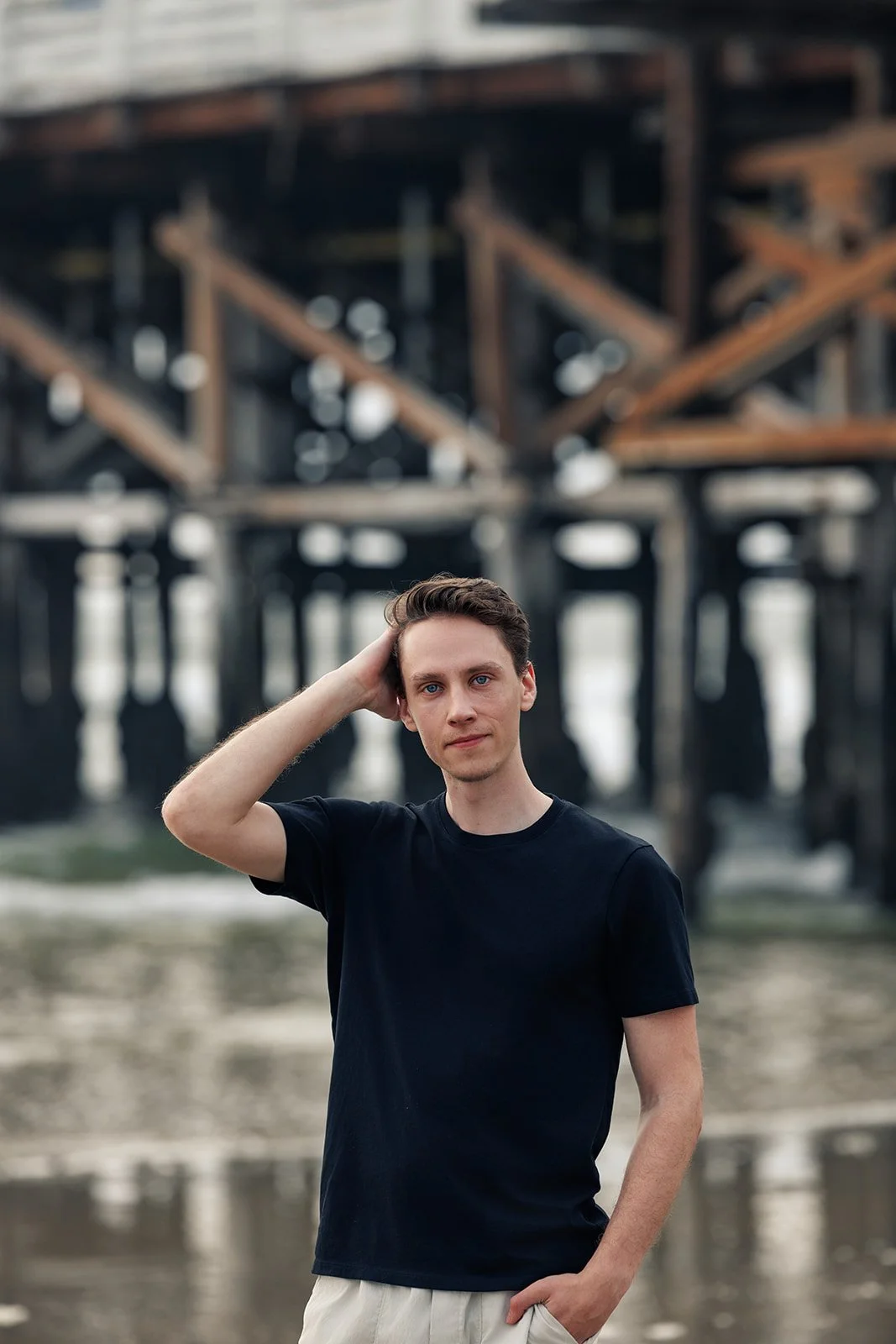 A young man stands outdoors near water with a large wooden pier in the background. He has short dark hair, blue eyes, and is wearing a black t-shirt and light-colored pants. He is touching his head with his right hand and looking at the camera.
