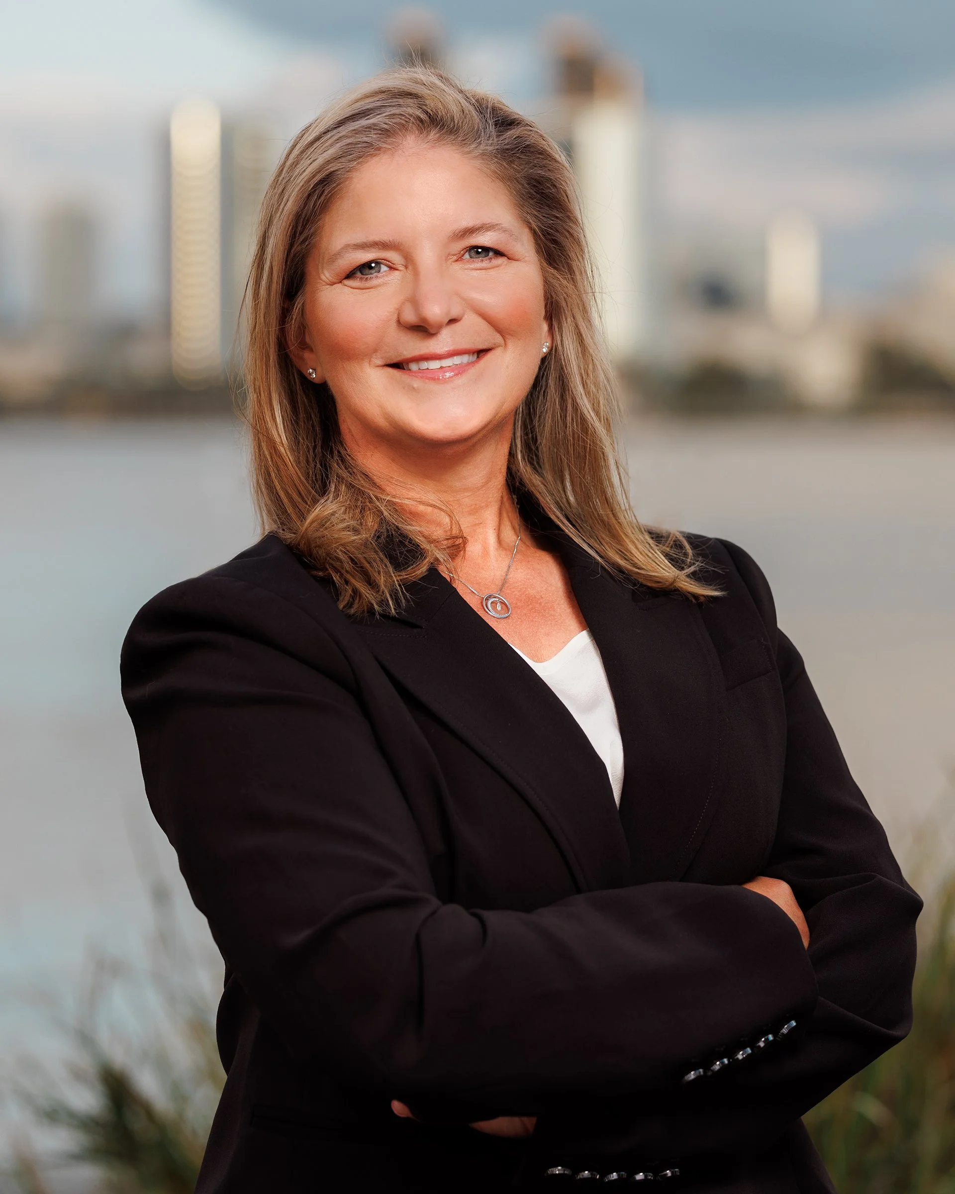 A woman in a black blazer with crossed arms smiling outdoors with a city skyline and water in the background.
