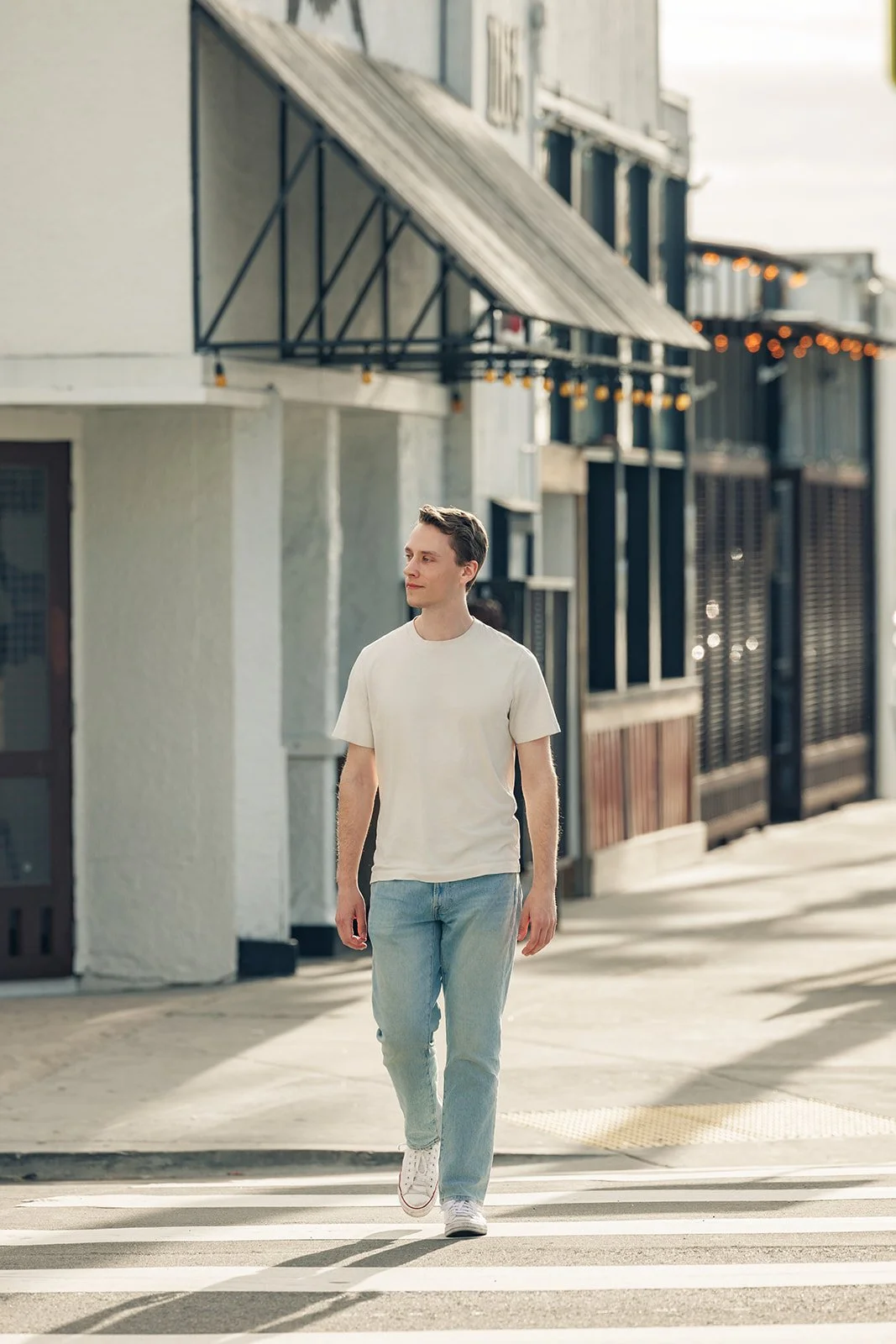 A young man walks across a city street crosswalk on a sunny day, wearing a white t-shirt, light blue jeans, and white sneakers.
