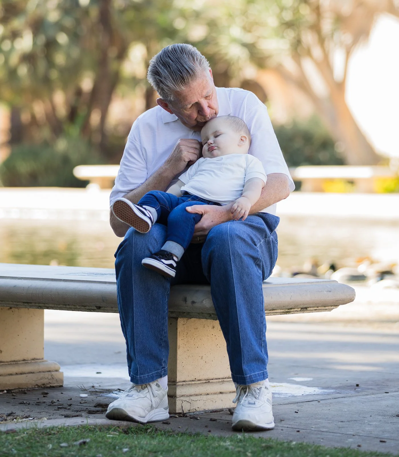 An elderly man with gray hair sitting on a park bench, holding a young child, possibly a toddler, who is sleeping with his head resting on the man's shoulder. The man is wearing a white t-shirt, blue jeans, and white sneakers. The background shows tr