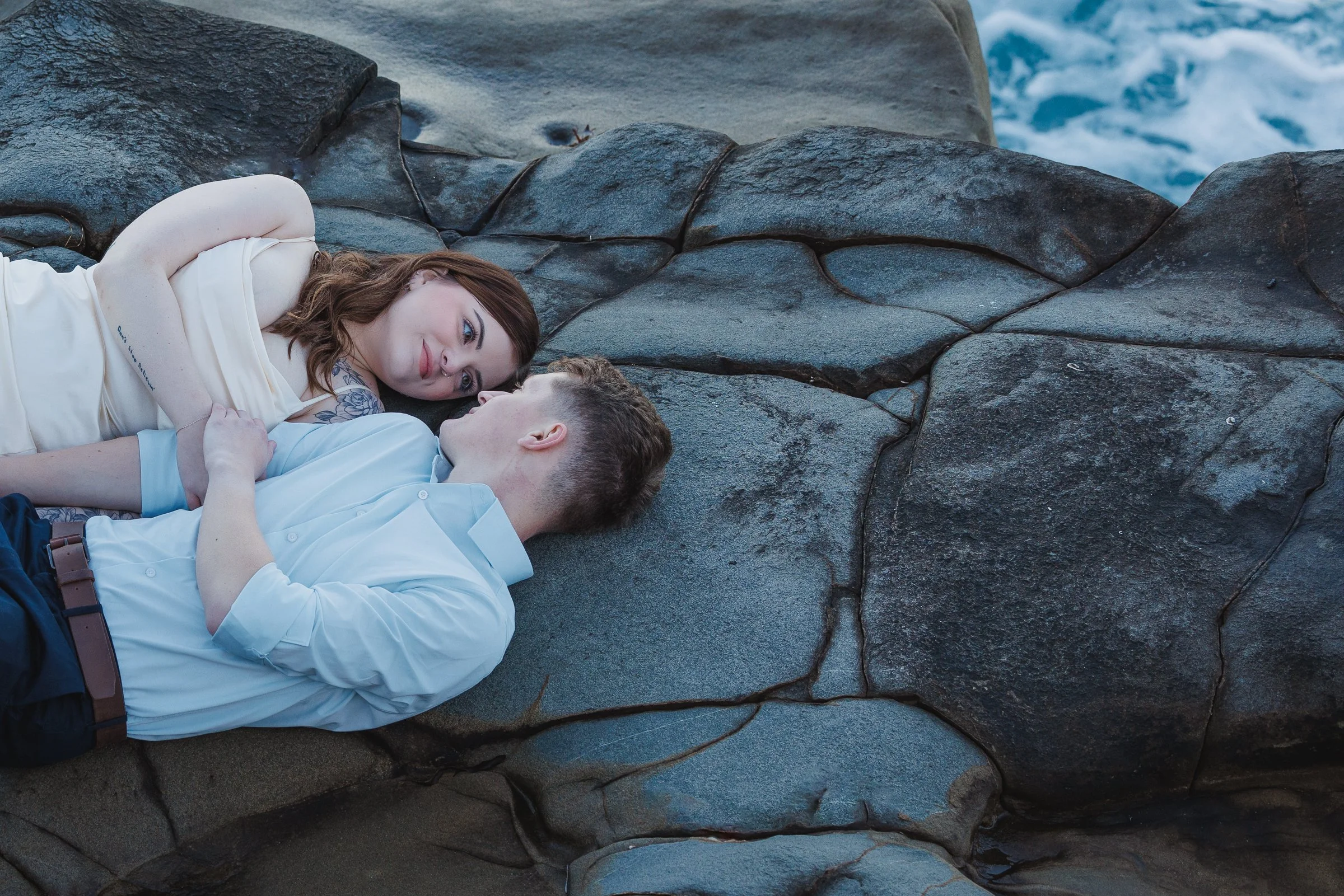 A man and woman lying on large rocks near the water, looking at each other affectionately.