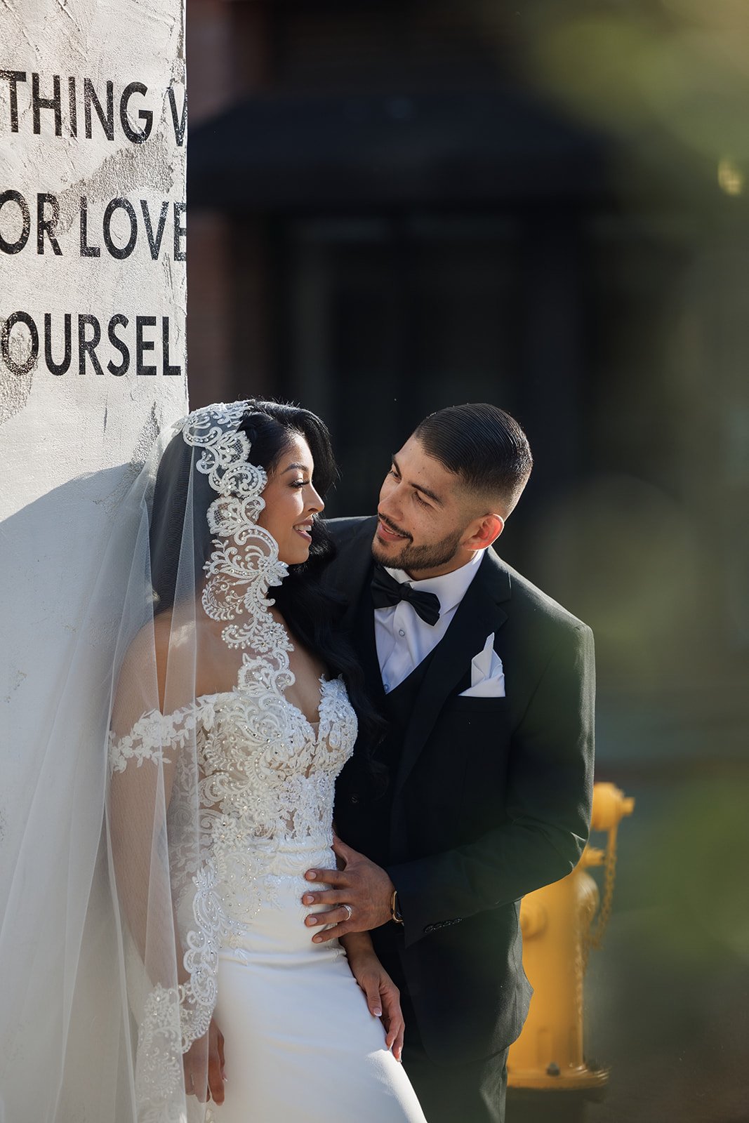 A newlywed couple dressed in wedding attire pose closely together outdoors, with the bride wearing a lace wedding dress and veil, and the groom in a black tuxedo with a bow tie, standing near a white wall with text and a yellow fire hydrant in the ba