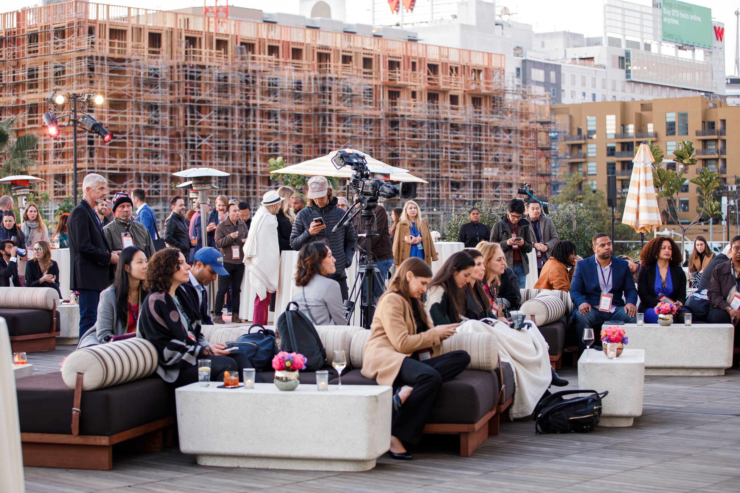 People attending an outdoor event on a rooftop, sitting on benches and sofas, with a construction site and high-rise buildings in the background. There are cameras, a photographer, and some individuals using their phones.