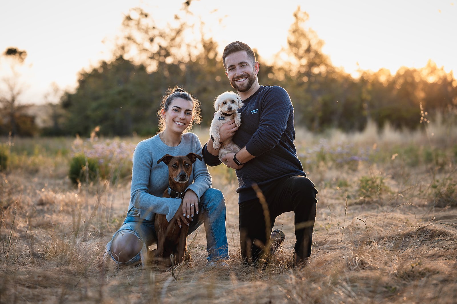 A smiling young woman and man kneeling on a field at sunset, each holding a dog, with dry grass and trees in the background.