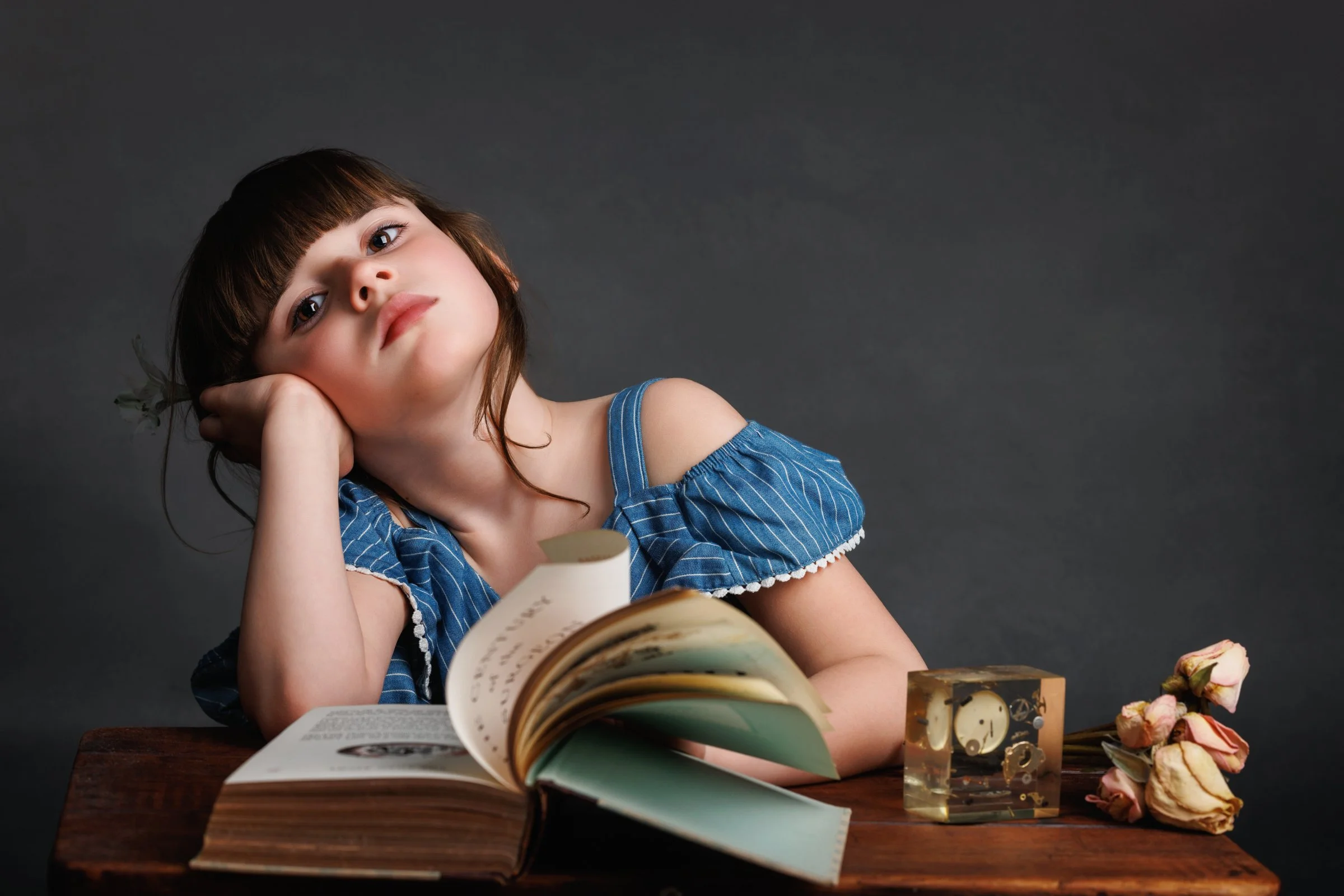 A young girl with brown hair and bangs, wearing a blue and white striped dress, rests her head on her left hand while sitting at a wooden table. In front of her is a partially open book, a small clock encased in a clear block, and a bunch of dried pi