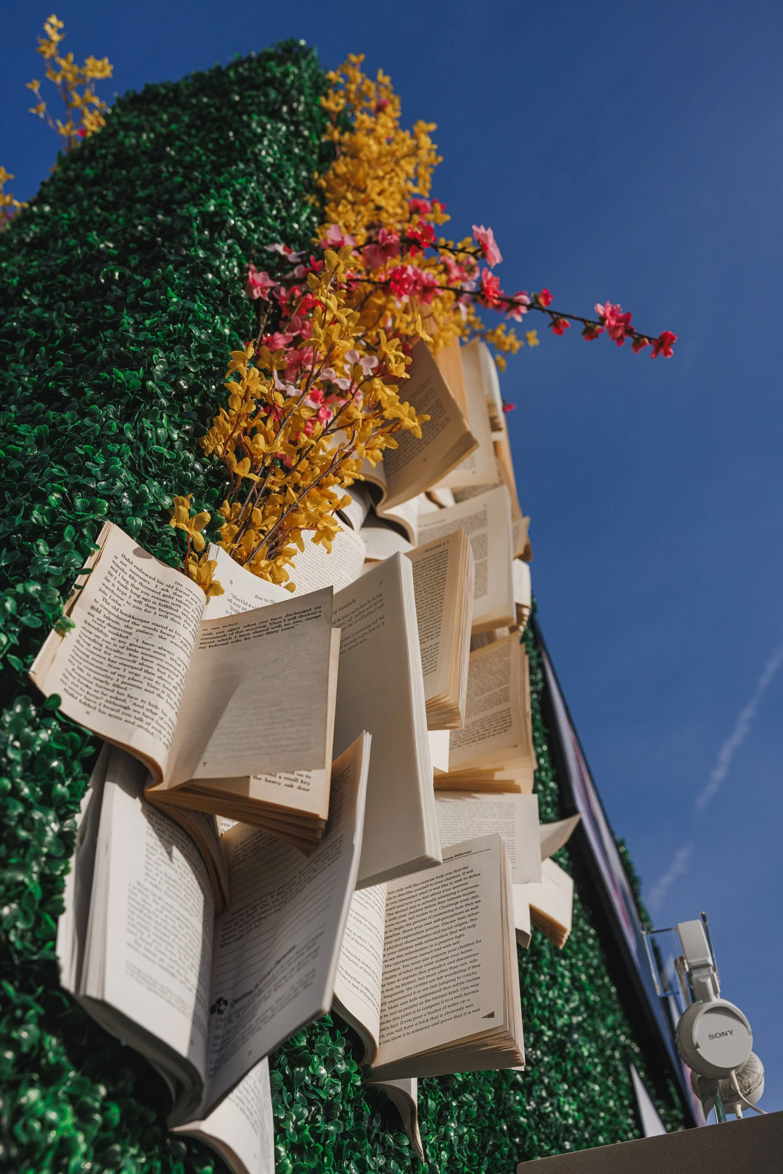 Open books arranged vertically on a green hedge with colorful artificial flowers and a clear blue sky in the background.