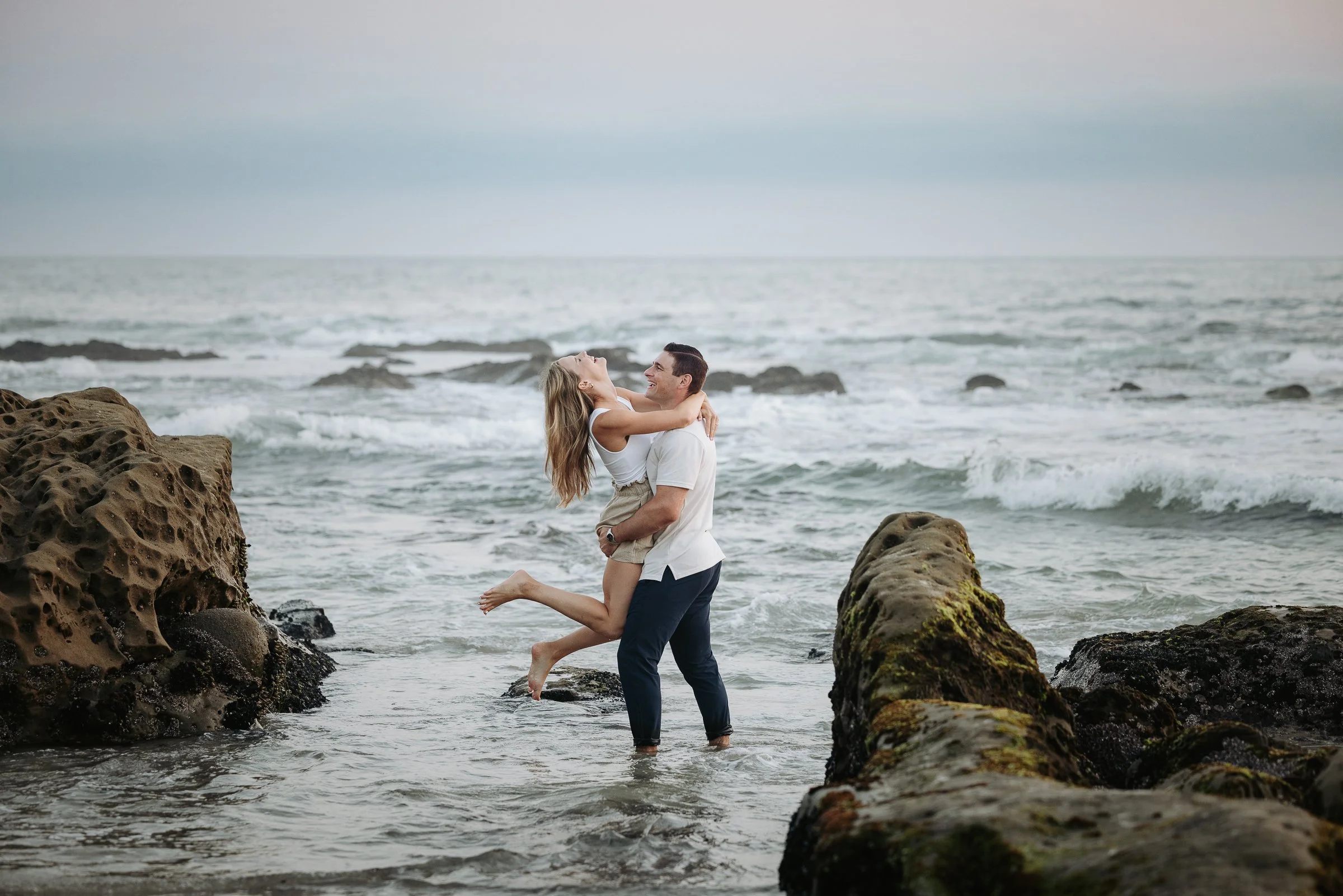 A couple at the beach, the man is lifting the woman who is smiling and embracing him, with rocks and the ocean in the background.