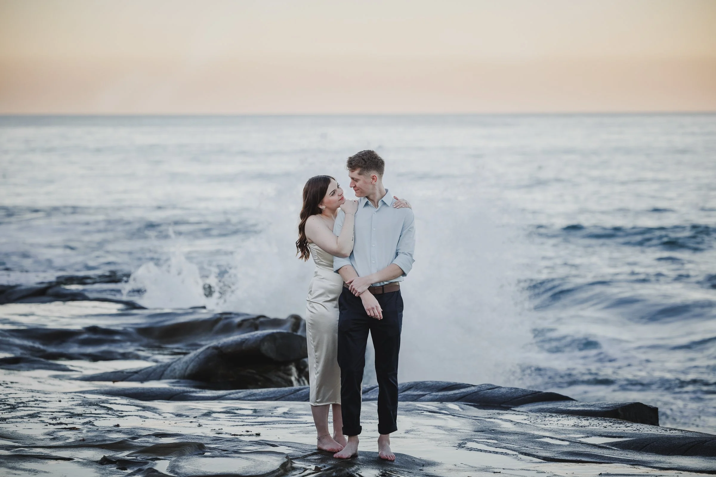 A couple standing on rocks by the ocean at sunset, gazing at each other.