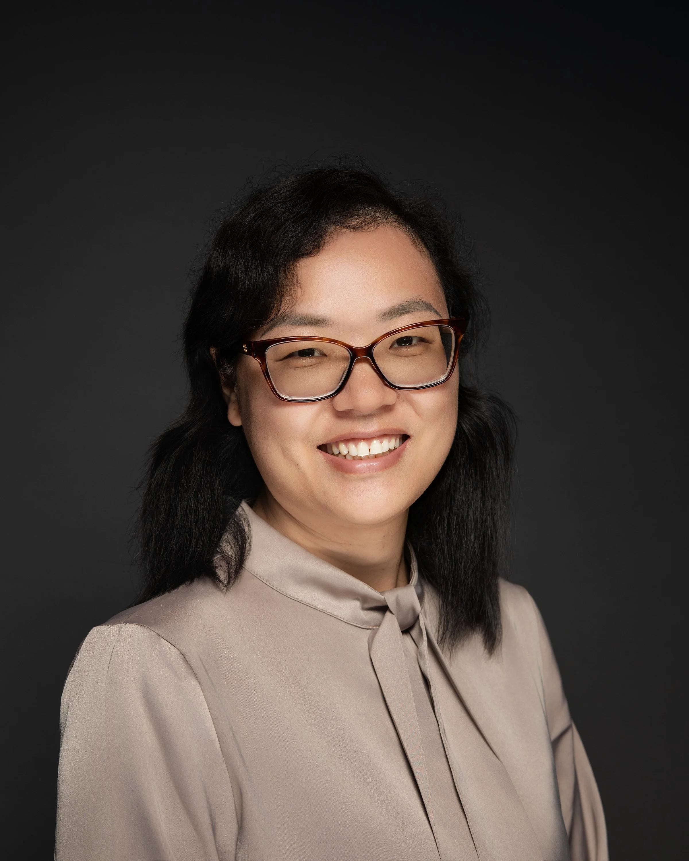 A woman with shoulder-length black hair, glasses, and a beige blouse smiling against a dark background.