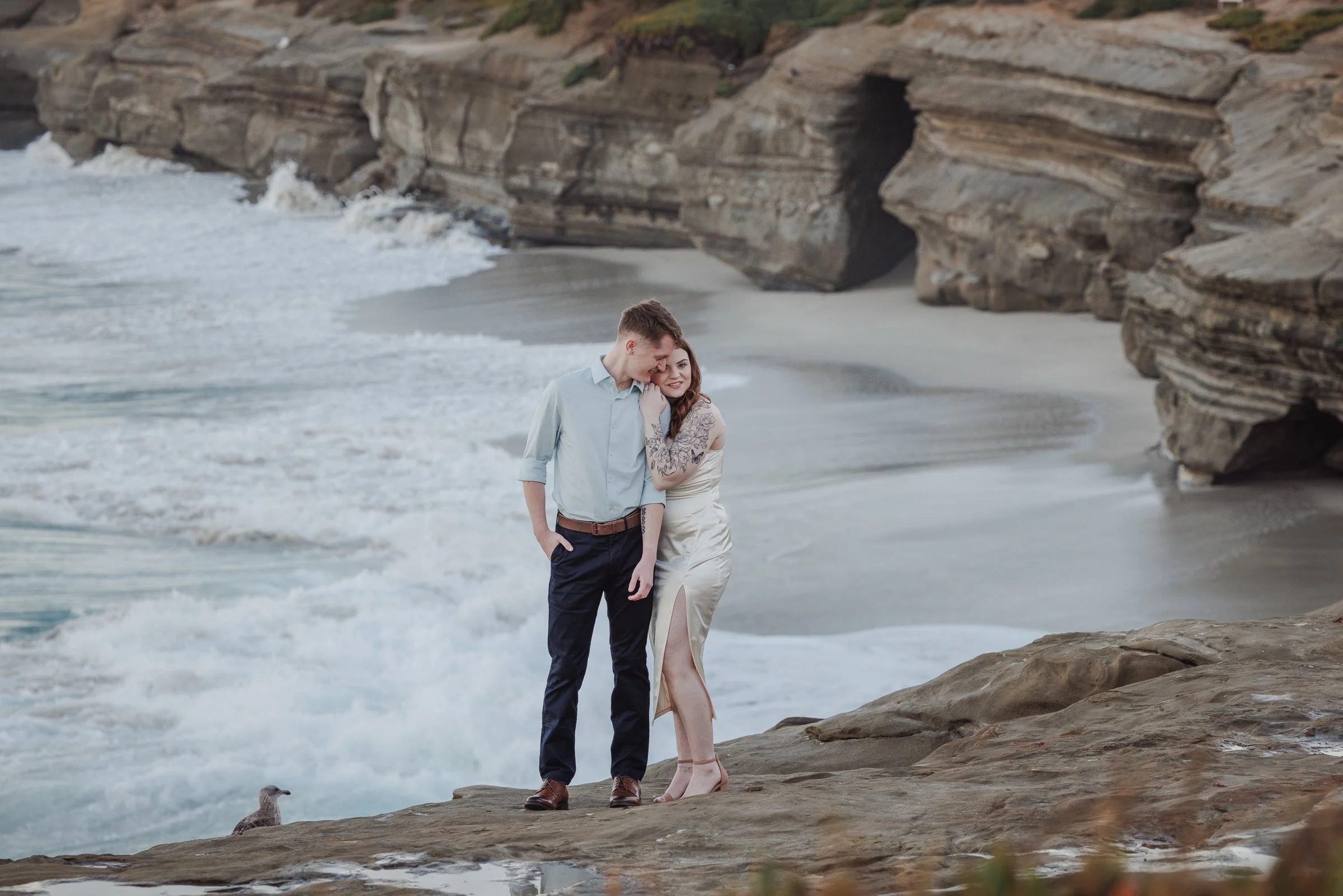 A couple standing on rocks near the beach, with waves and cliffs in the background, sharing a tender moment as they smile and lean into each other.