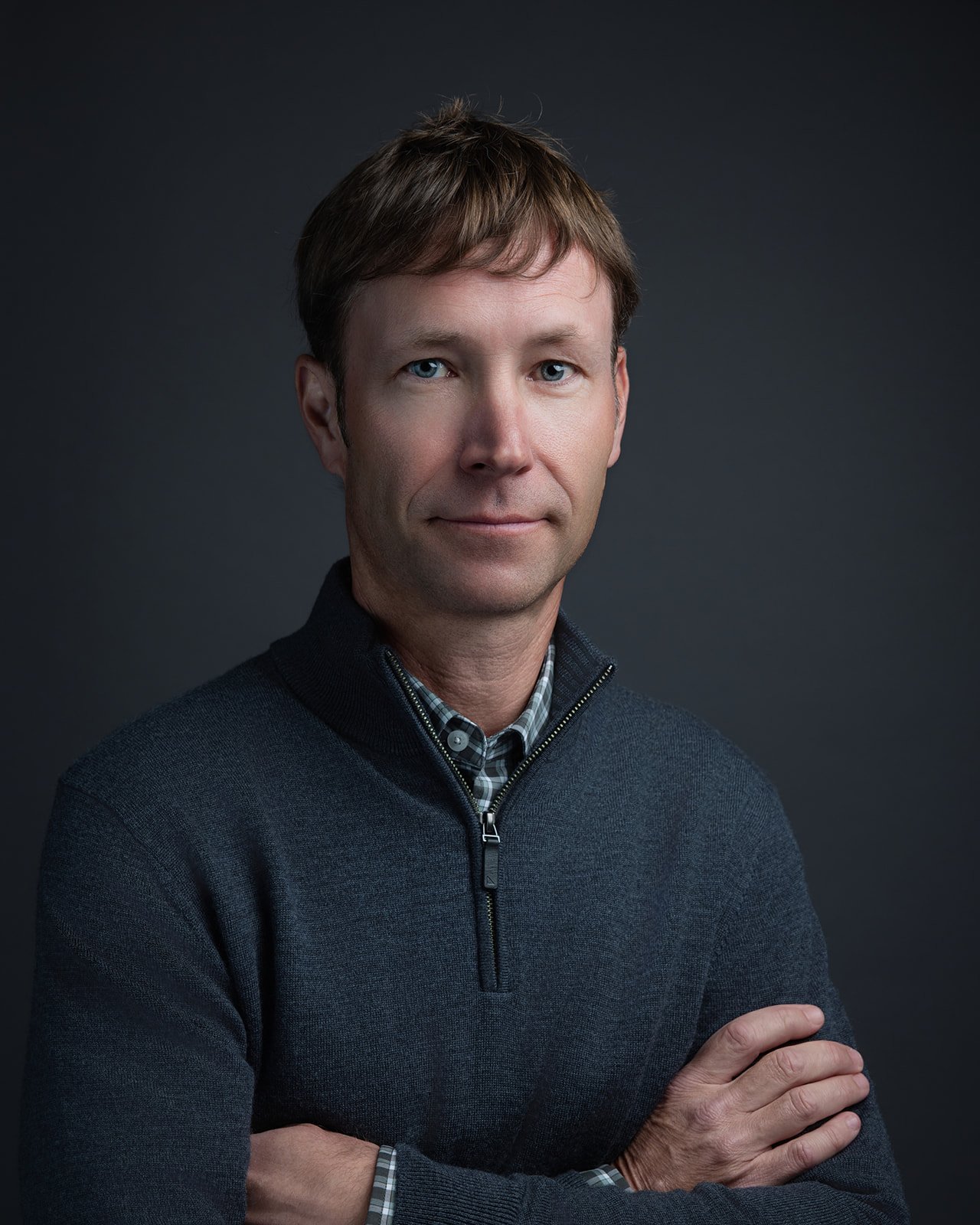 A portrait of a man with light brown hair, blue eyes, and fair skin, wearing a dark gray sweater over a checkered shirt, with arms crossed against a dark background.