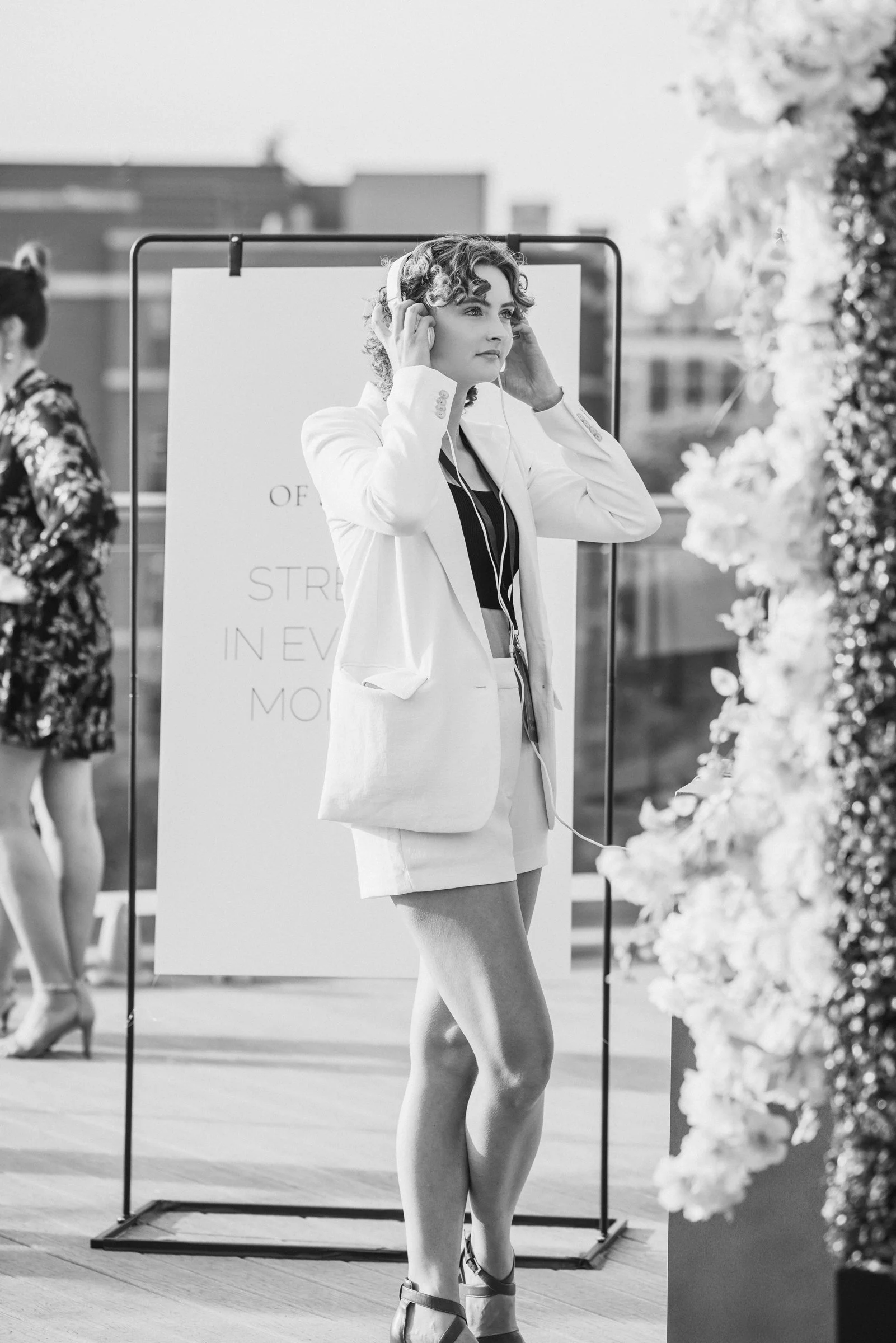 A woman with curly hair listens to music with headphones on, standing outdoors near floral decorations, on a city street with a sign in the background.
