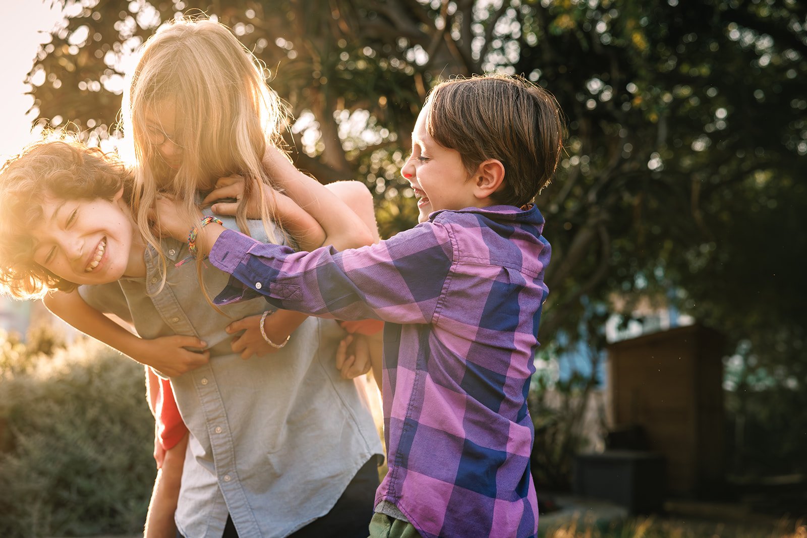 Three children having a playful fight outdoors, smiling and laughing, with sunlight filtering through trees in the background.
