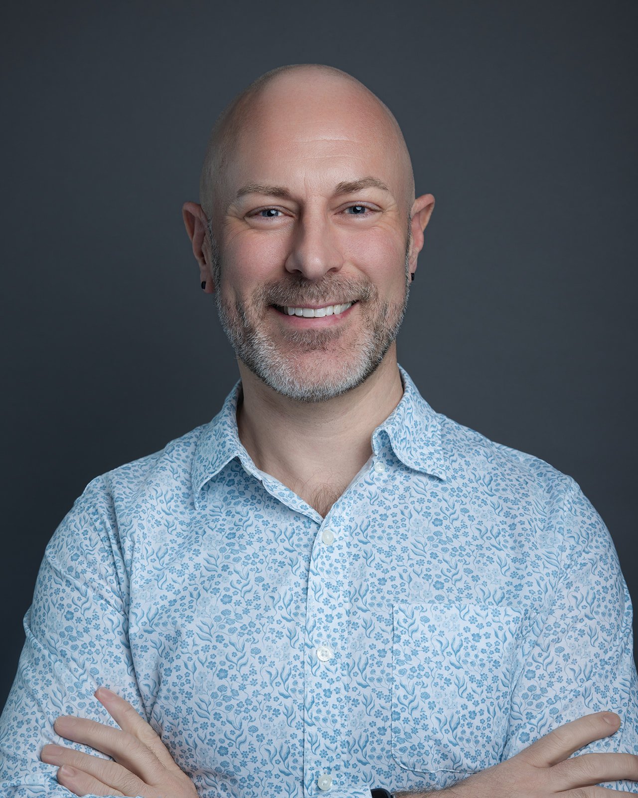A smiling man with a bald head, beard, and earrings, wearing a patterned white and blue shirt, against a dark background.