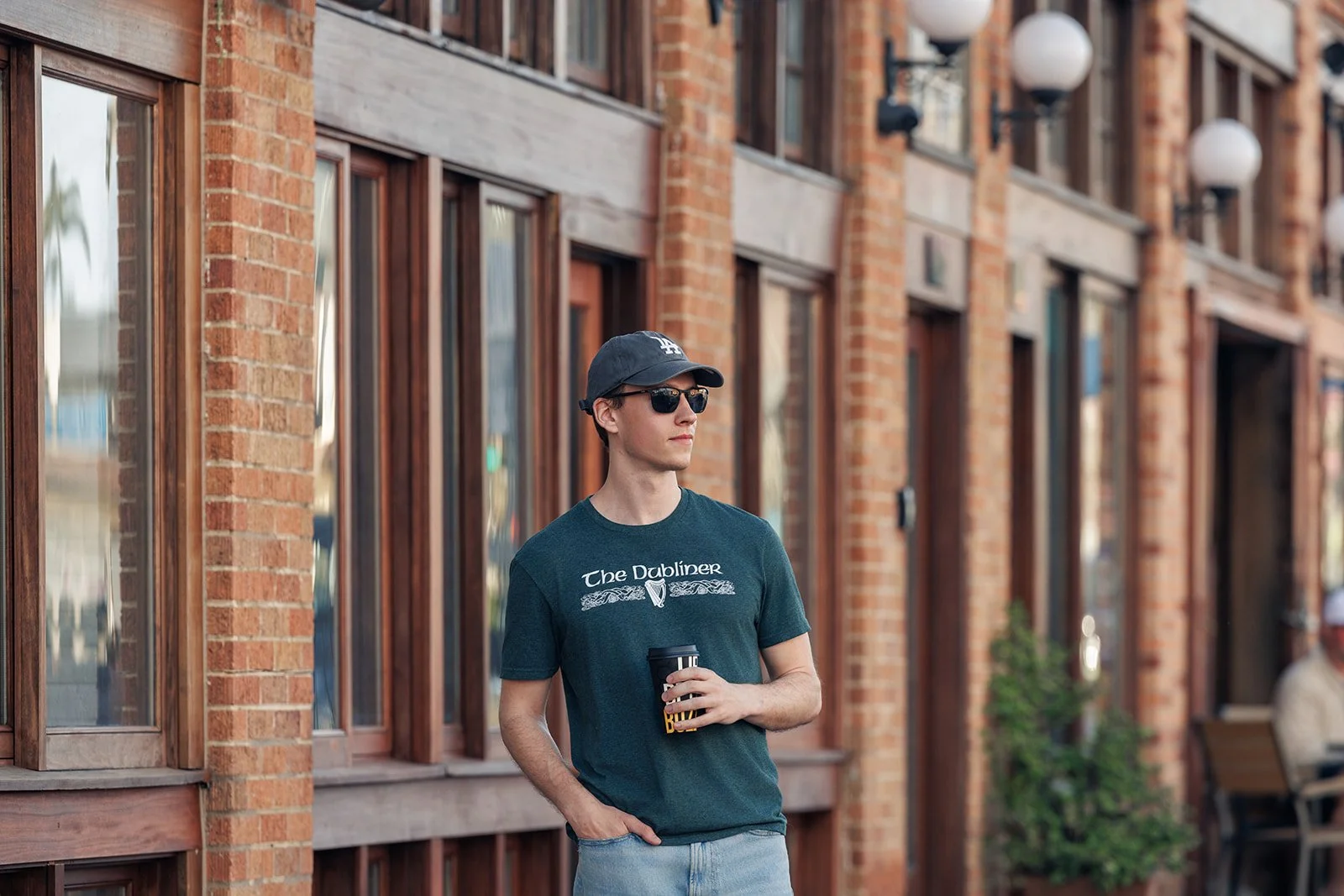A young man wearing a navy cap, sunglasses, a dark green T-shirt, and jeans, holding a to-go coffee cup, standing outdoors in front of a brick building with wooden framed windows.