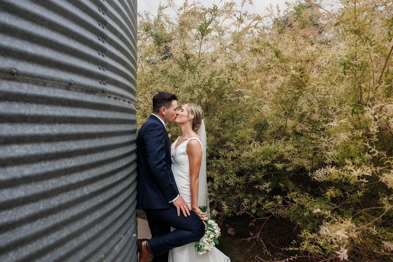 A newlywed couple sharing a kiss outdoors, with the groom leaning against a large metal silo and surrounded by greenery.
