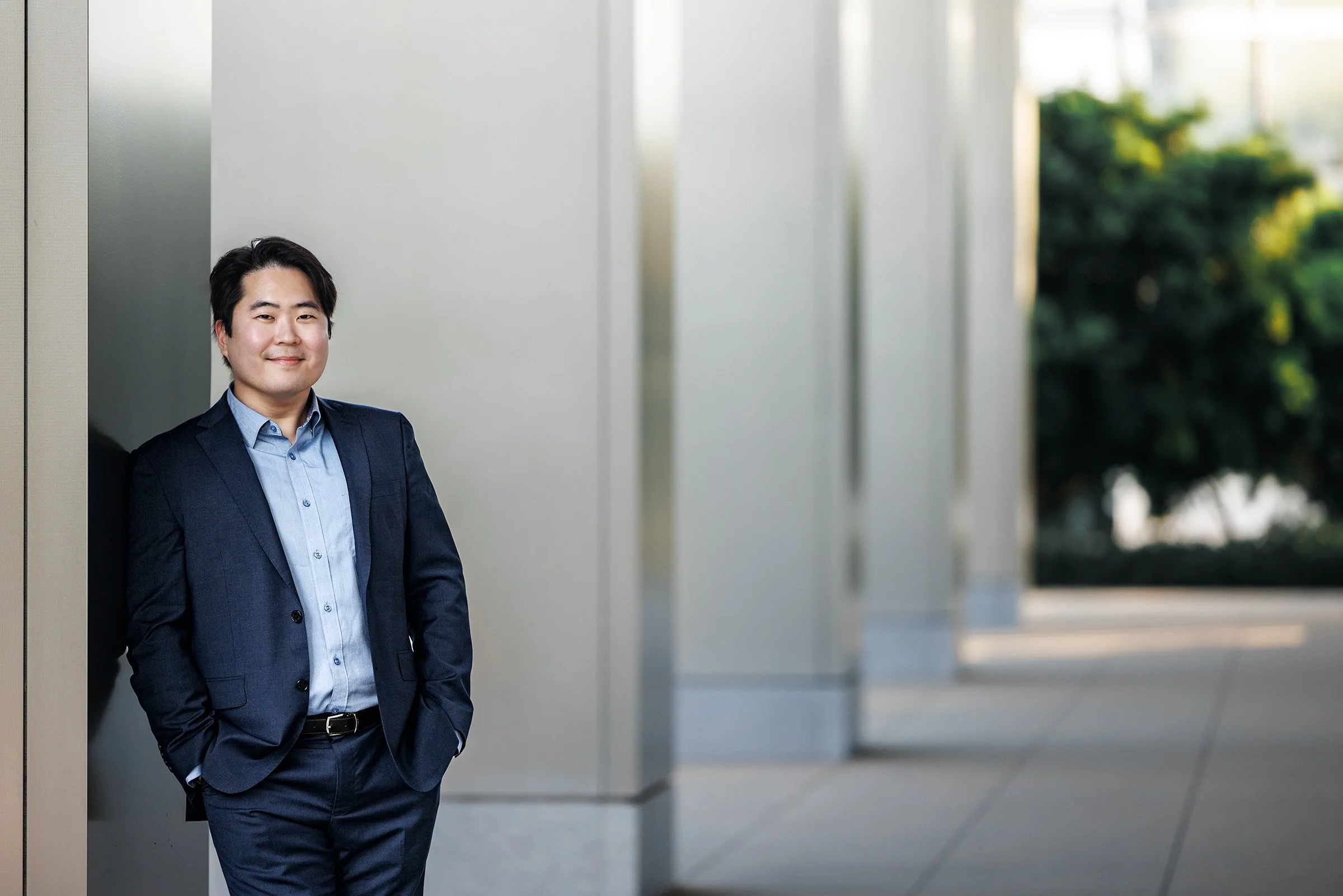 A young man in a navy blue suit and light blue shirt stands outdoors leaning against a wall, with a background of blurred trees and building columns.