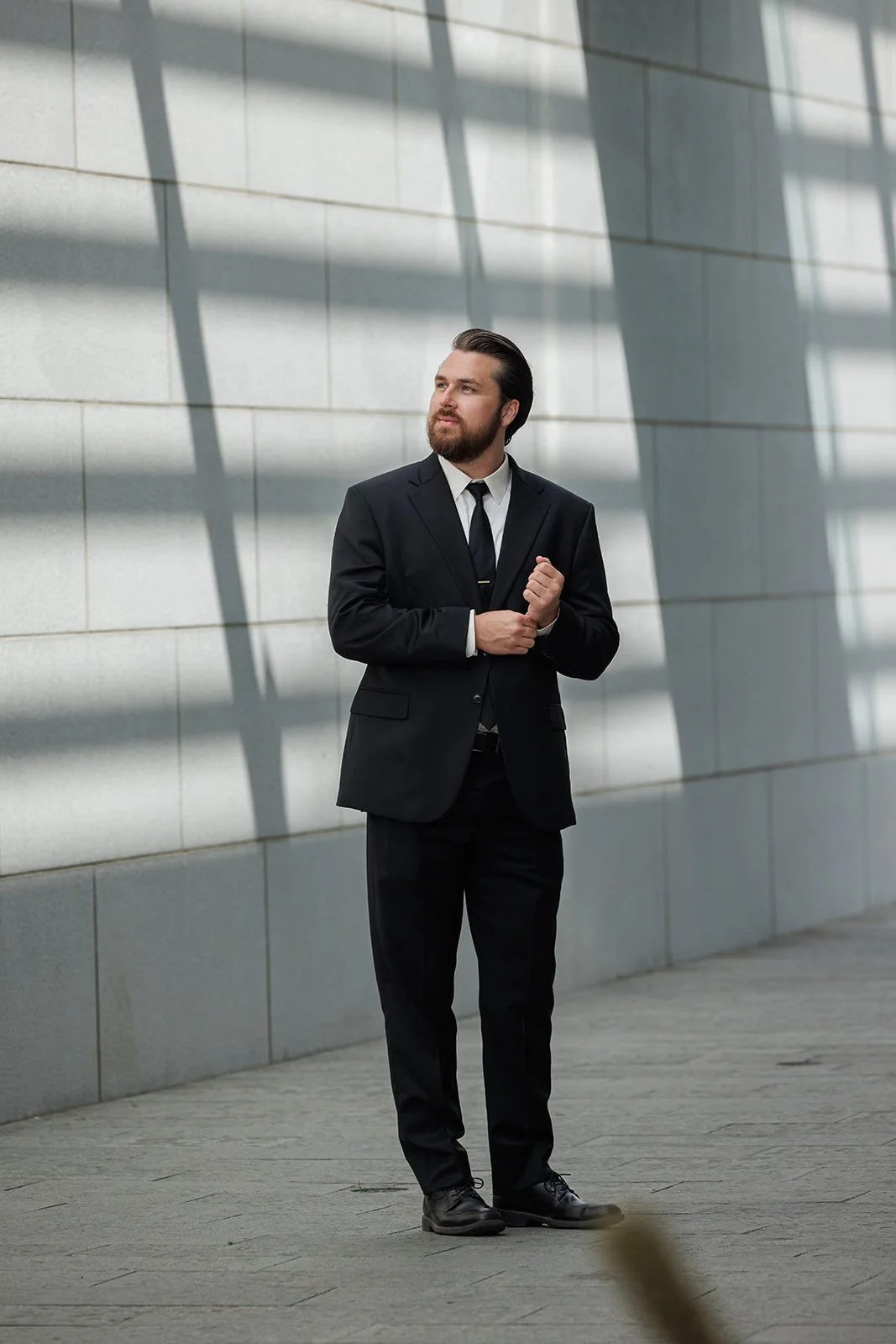 A man in a black suit, white shirt, and black tie standing outside against a stone wall with large window shadows, looking thoughtfully into the distance.