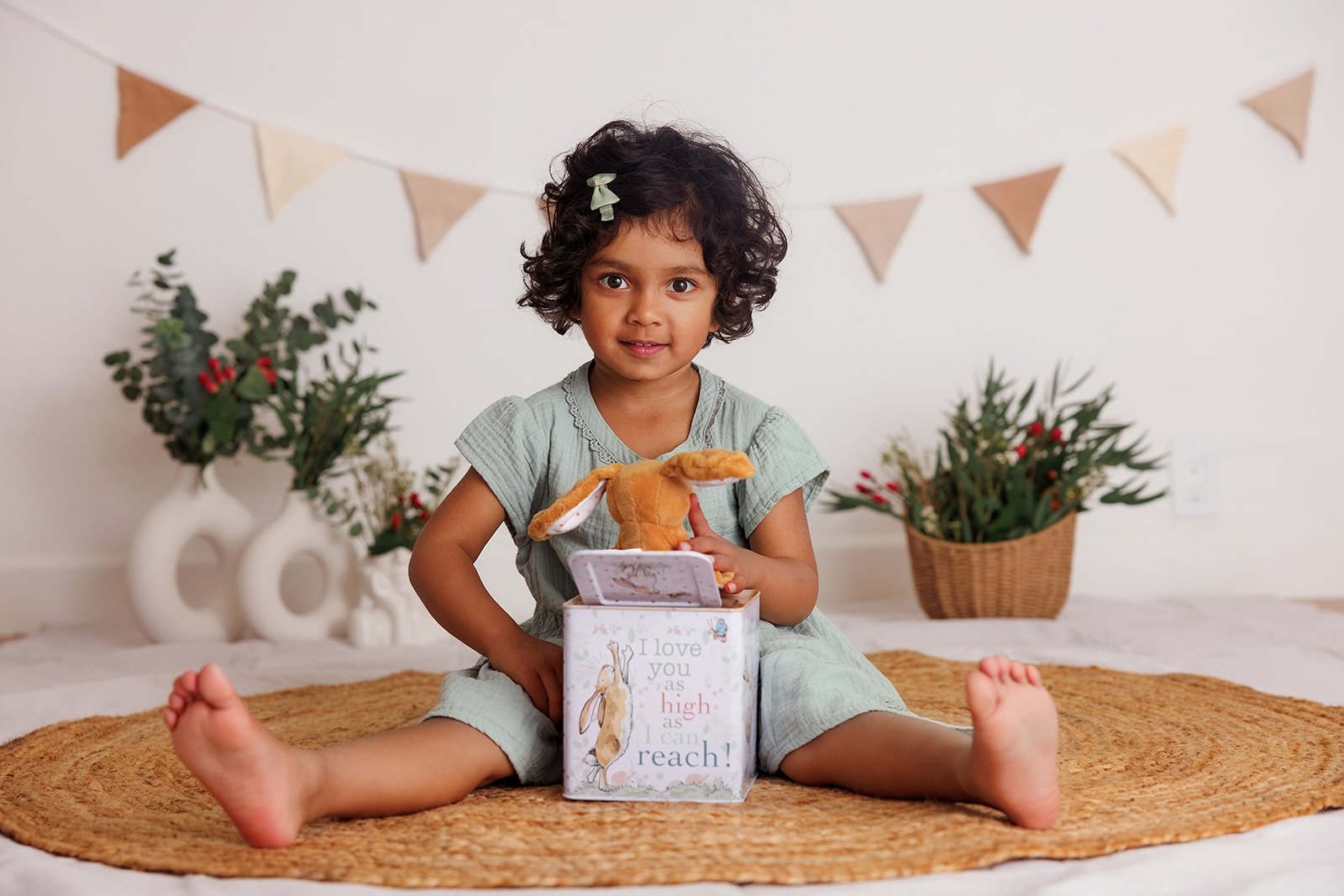 A young girl with curly hair and a green bow, sitting cross-legged on a woven rug, holding a stuffed bunny and a gift box with a bunny illustration, in a decorated room with plants and a banner.