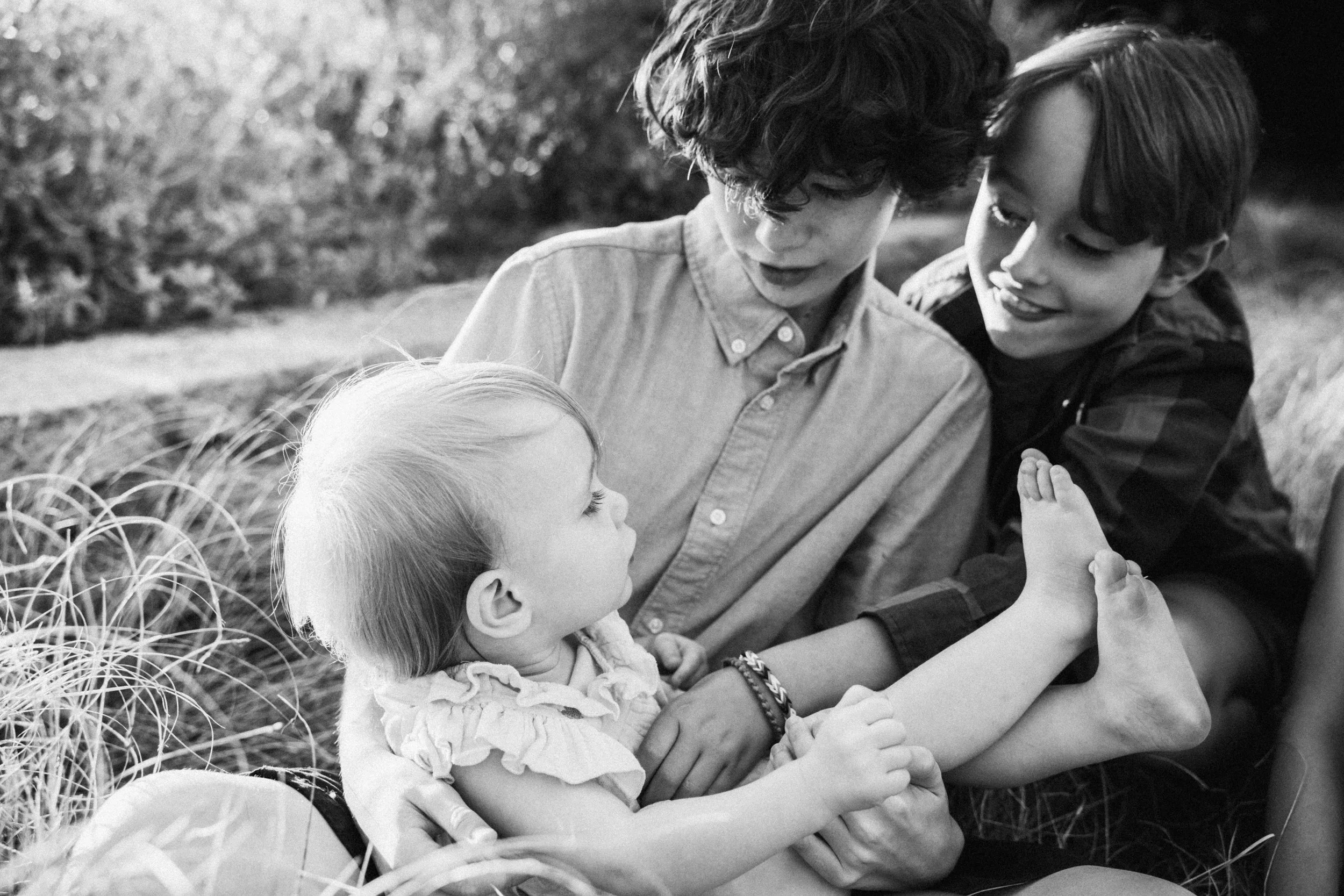A black-and-white photo of two boys and a young girl on a grassy area outdoors. The girl is lying down, holding her legs, while one boy supports her and the other boy watches with a smile.