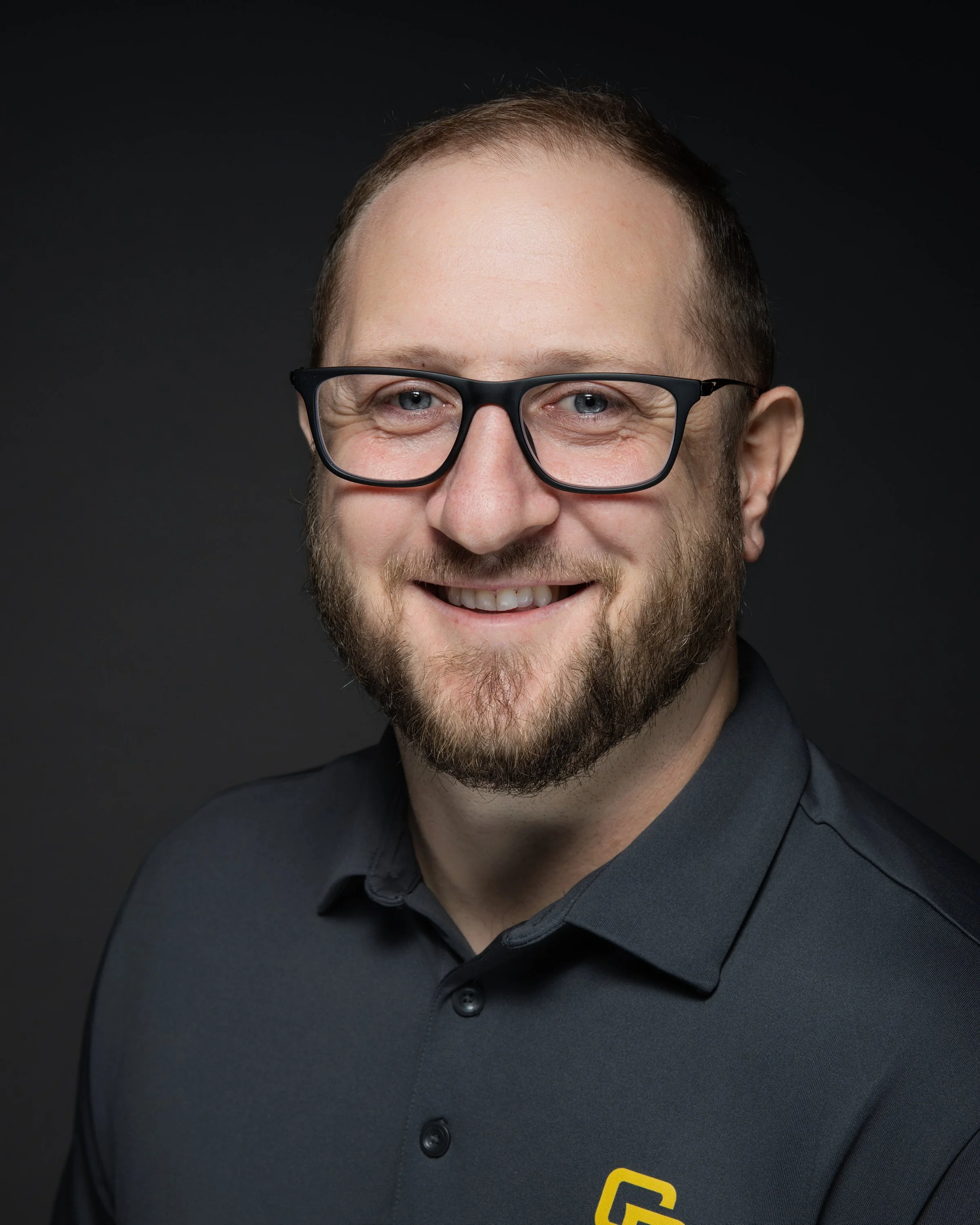 A smiling man with short hair, glasses, a beard, and a grey collared shirt with a yellow logo, against a dark background.