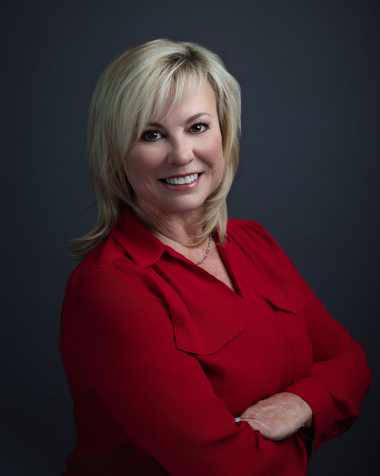Professional woman with blonde hair wearing a red blouse against a dark gray background.