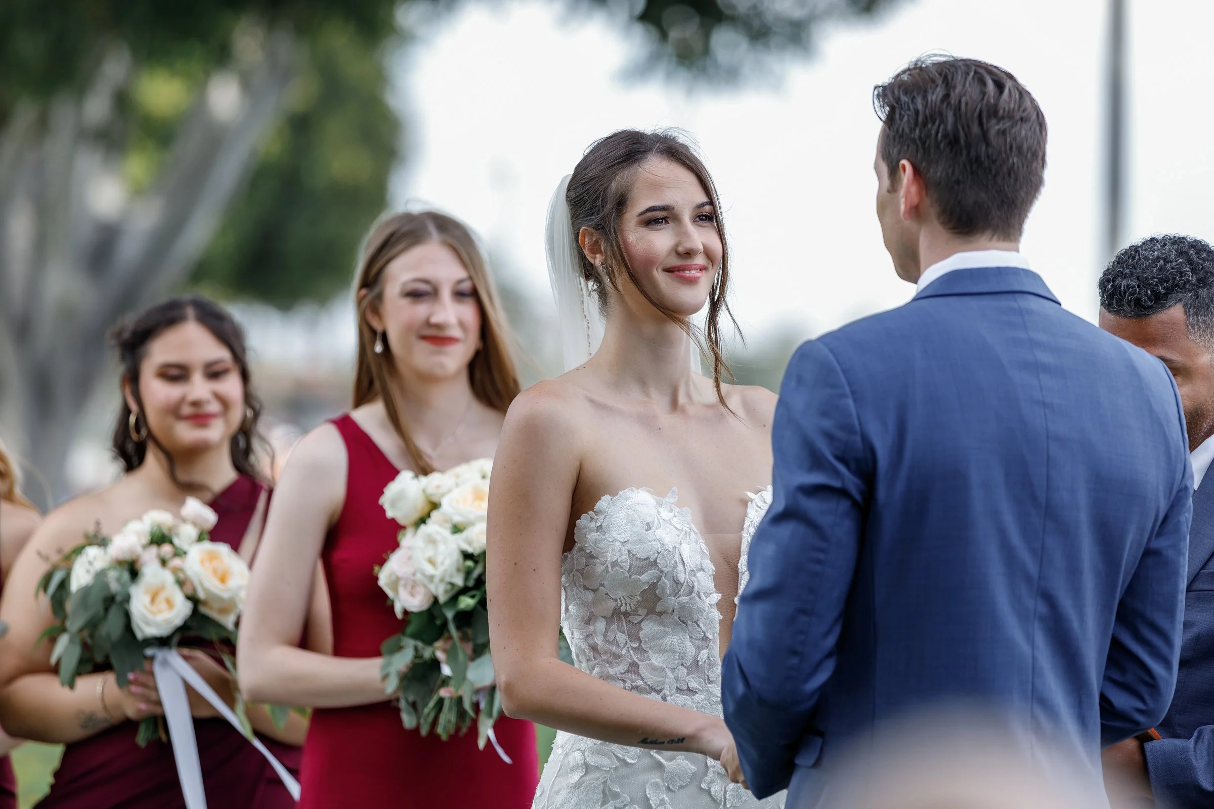 Bride and groom exchanging vows during outdoor wedding ceremony, surrounded by bridesmaids holding bouquets.
