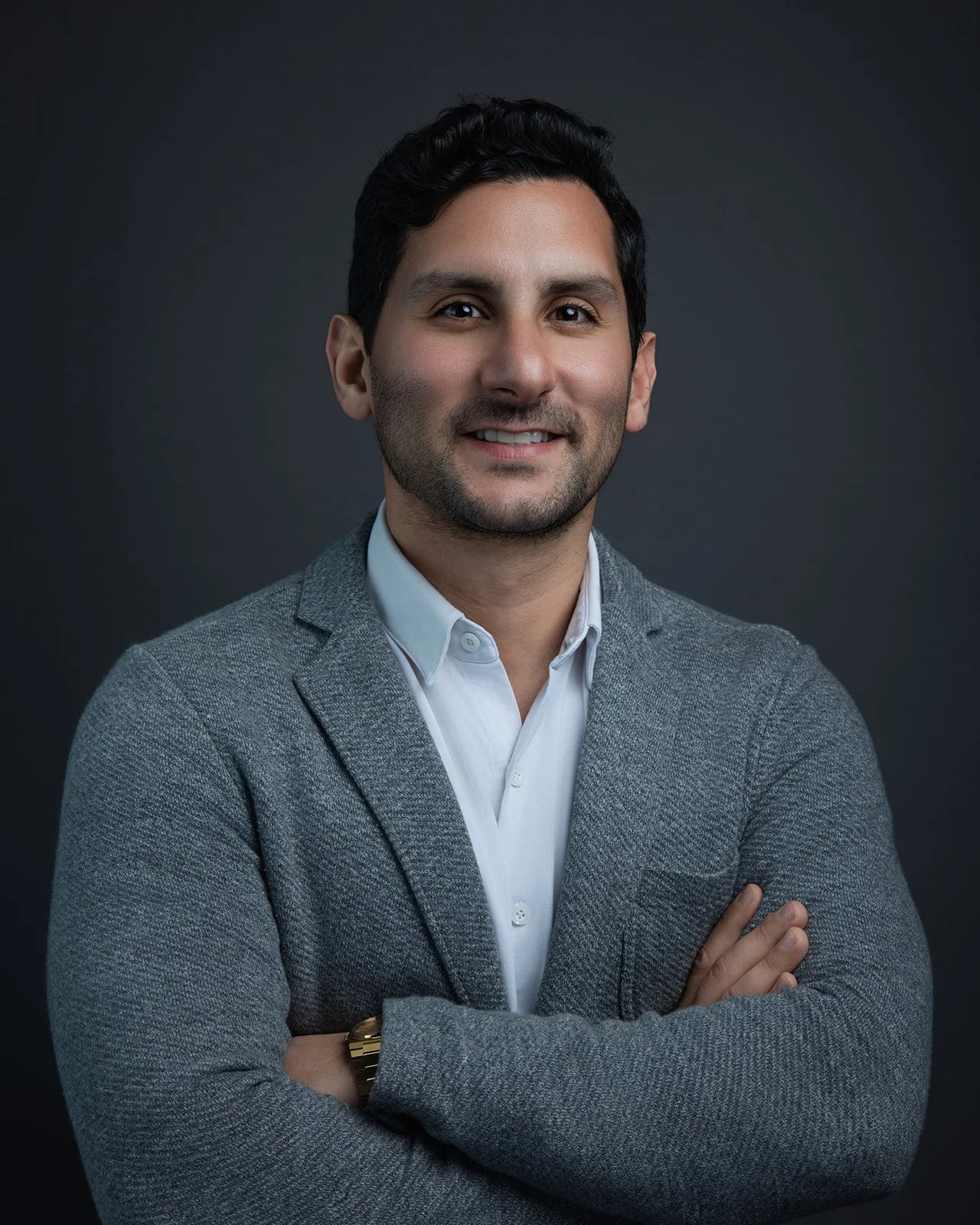 Headshot of a smiling man with dark hair, wearing a white shirt and gray blazer, crossing arms in front of a dark background.