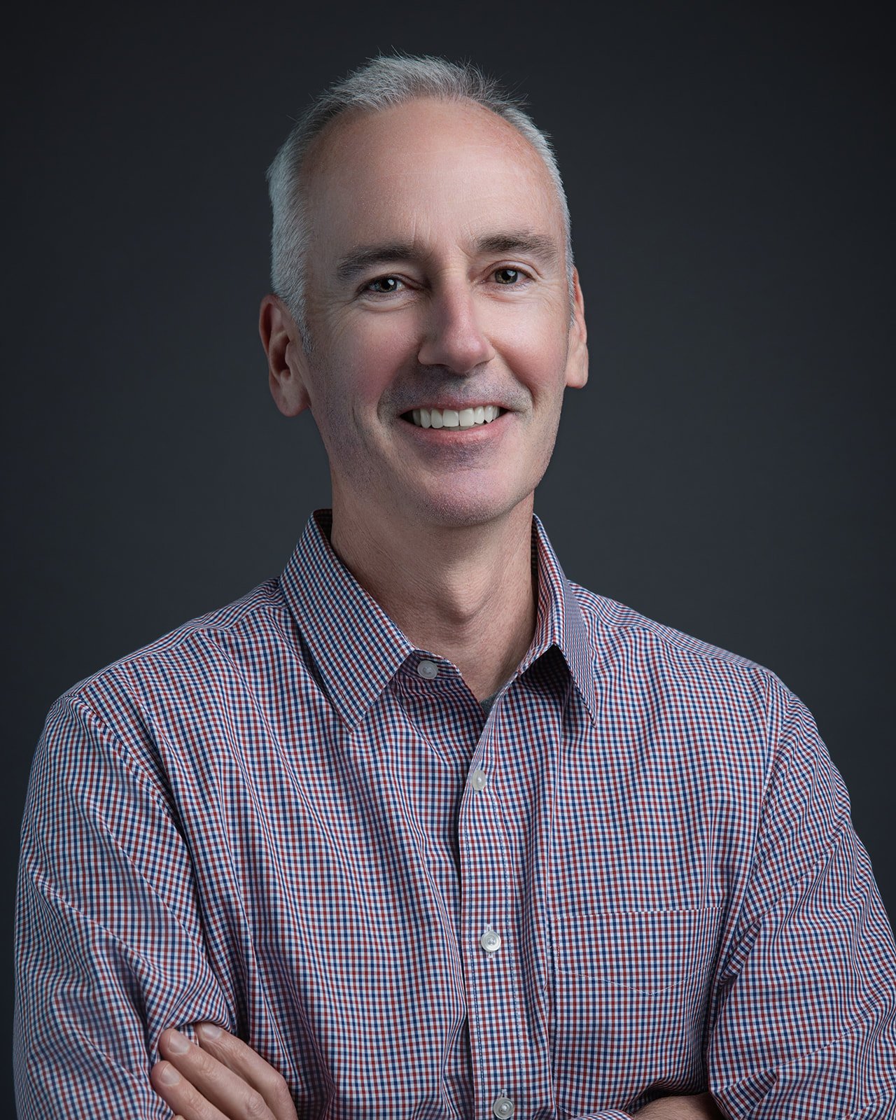 Portrait of a middle-aged man with gray hair, wearing a checkered shirt, smiling with arms crossed, against a dark background.