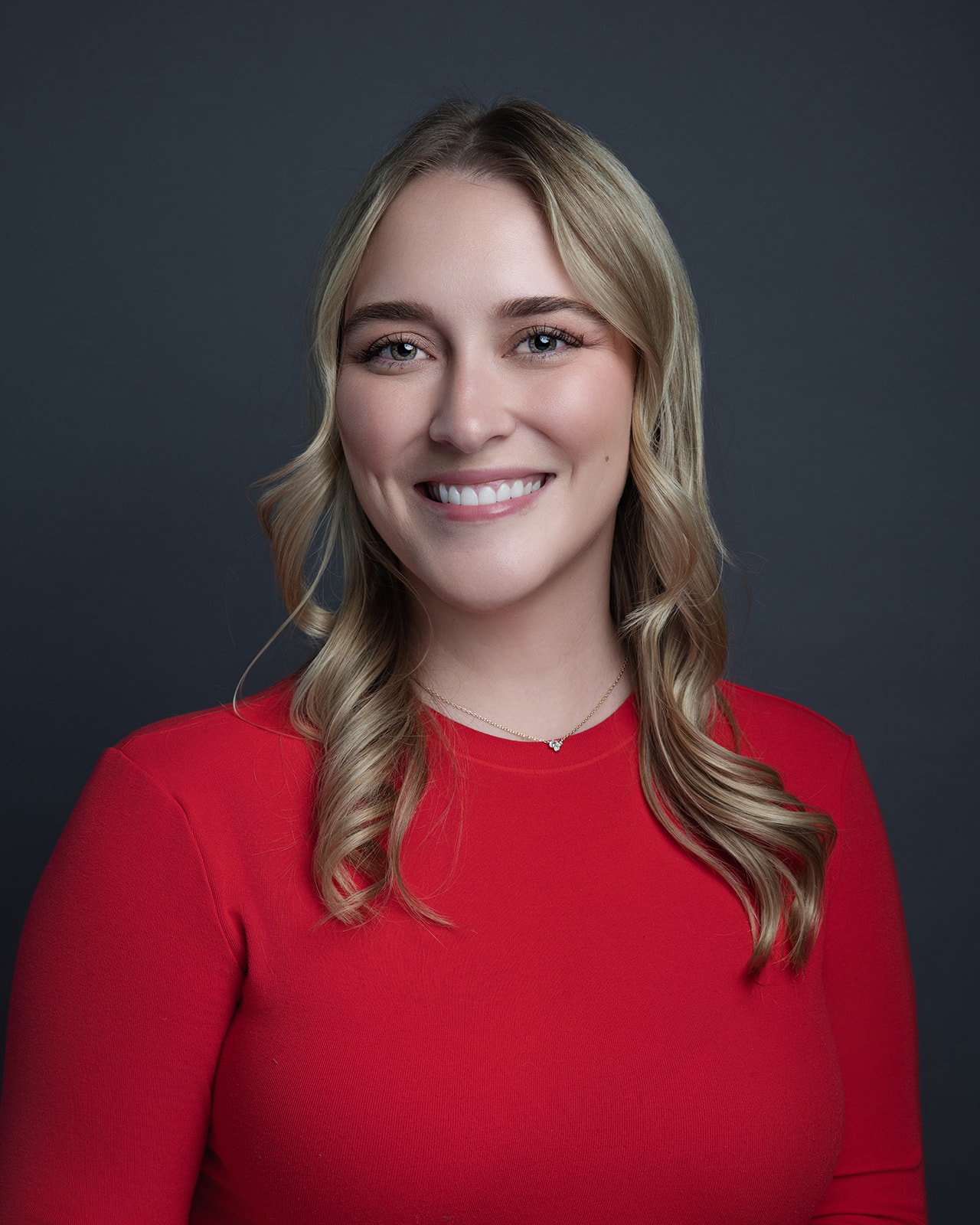 A smiling woman with wavy blonde hair wearing a red top and a delicate necklace, posing against a dark gray background.