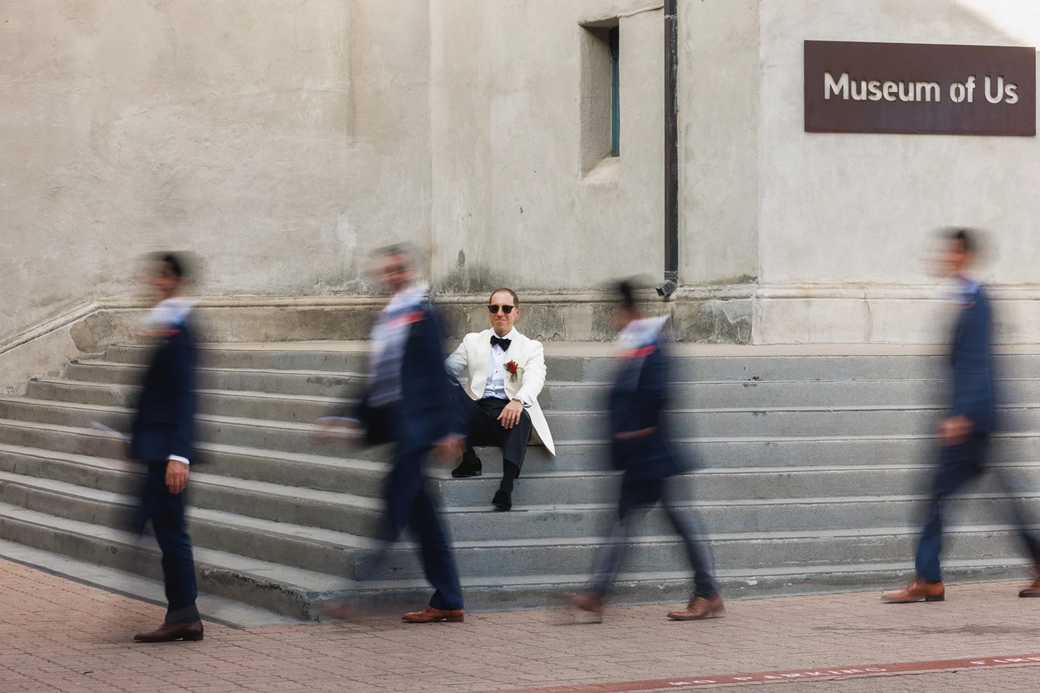 Groom seated on museum steps as groomsmen blur past