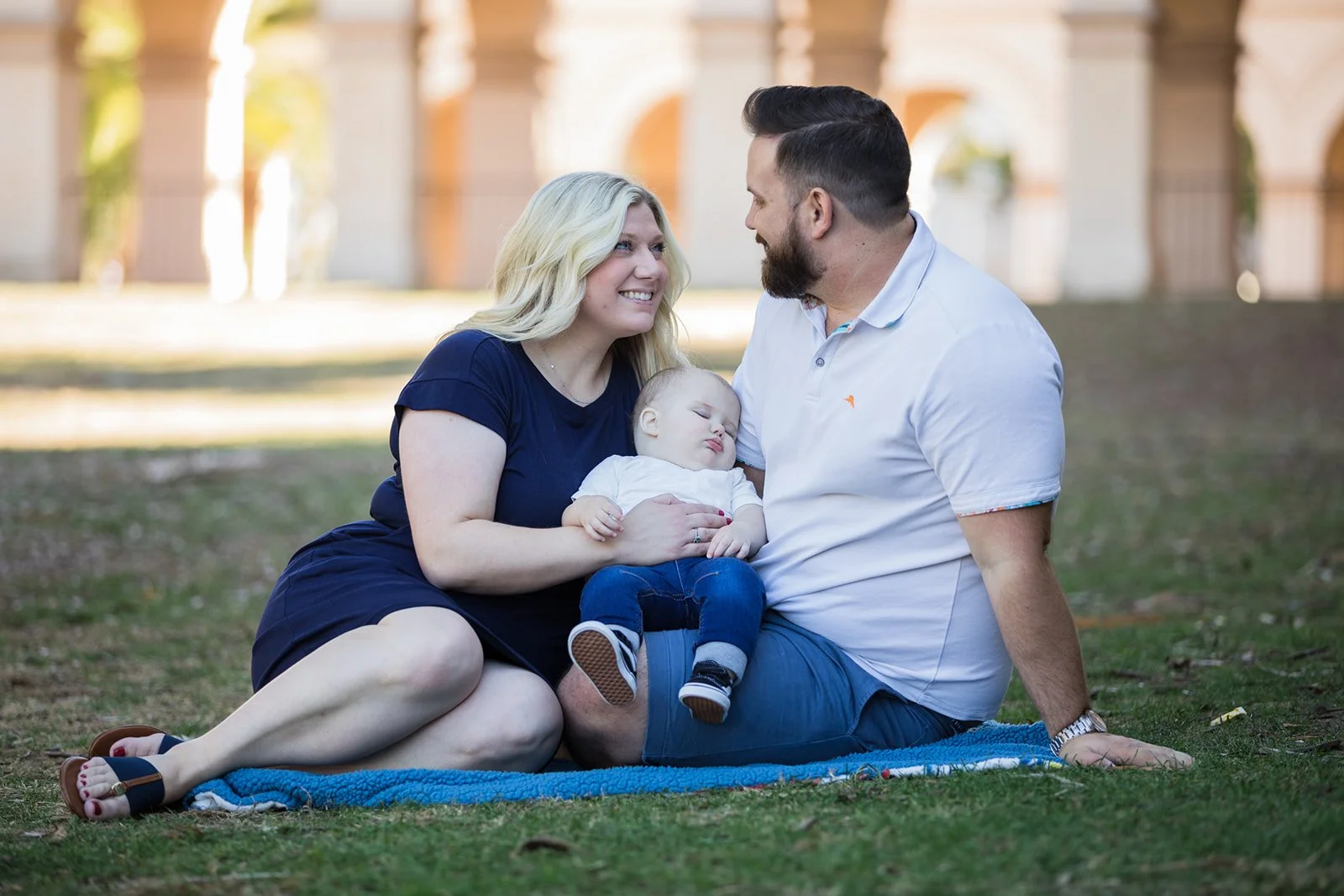 A family of three sitting on a blanket on the grass in a park, with a woman holding a sleeping child and a man looking at her, smiling.