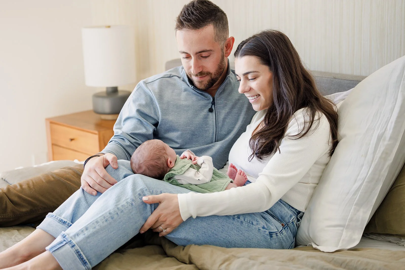 A family of three sitting on a bed, a woman holding a newborn baby with a man leaning over them, all smiling and looking at the baby.