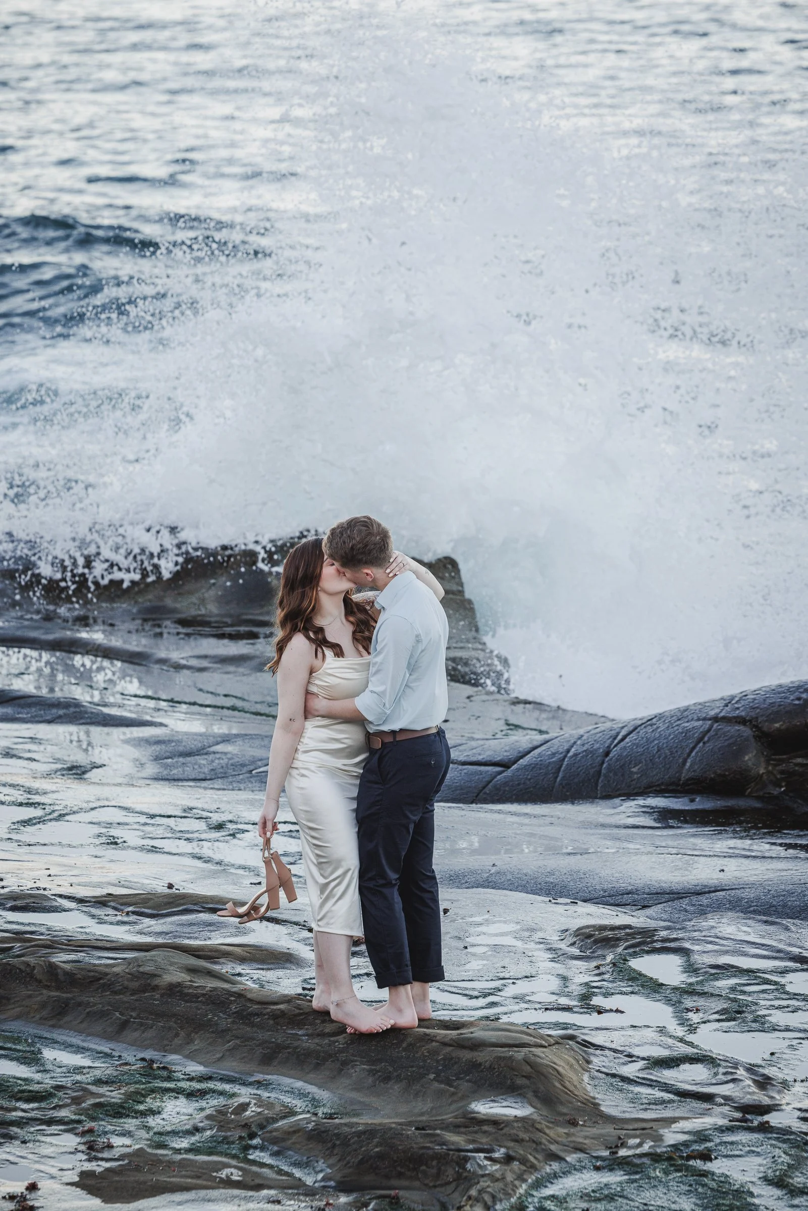 A couple kissing on a rocky beach with waves crashing behind them.
